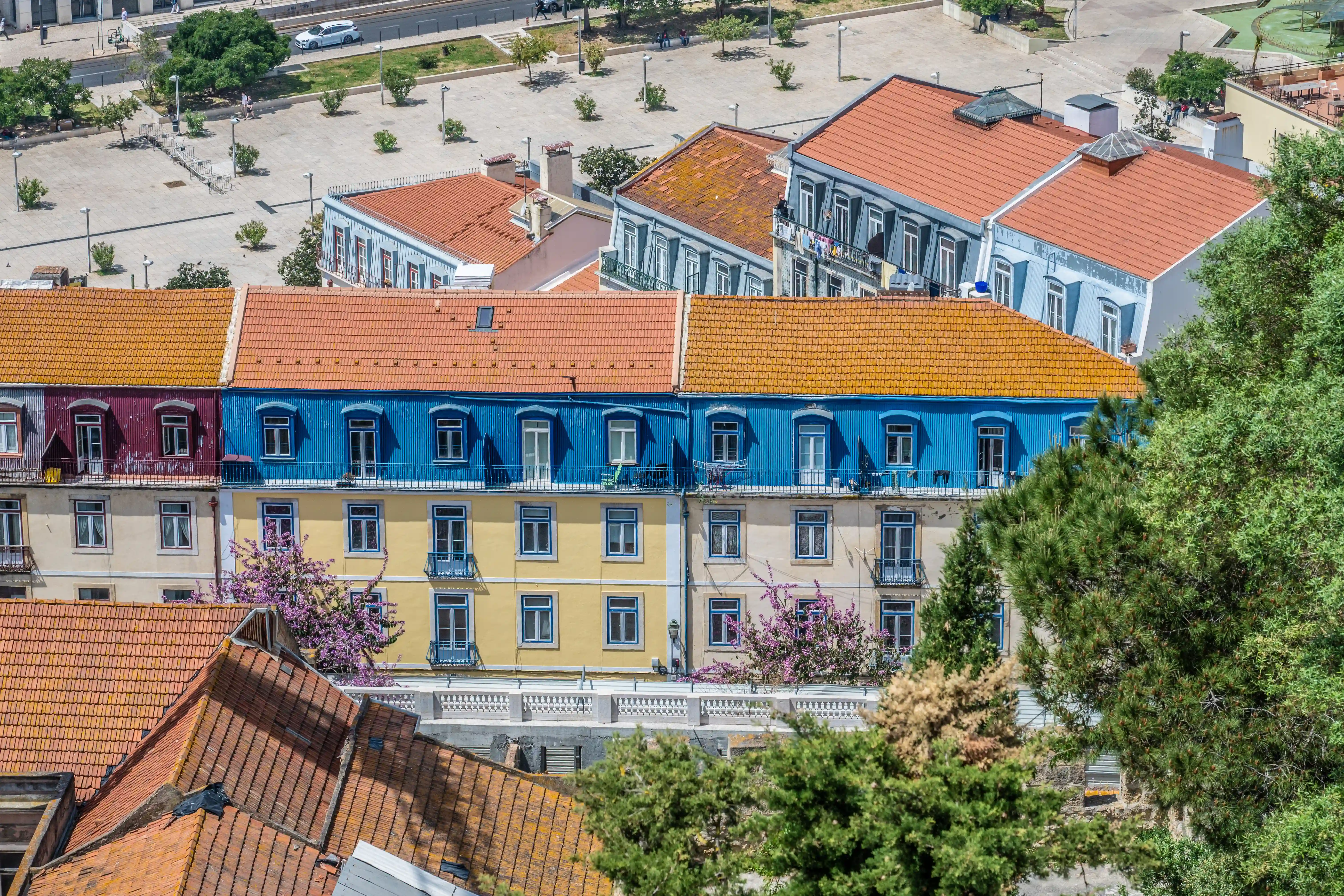 Aerial view of colourful apartment buildings in Lisbon with orange tiled roofs, blue and yellow facades, balconies, and trees with purple blossoms, overlooking a city plaza and street.