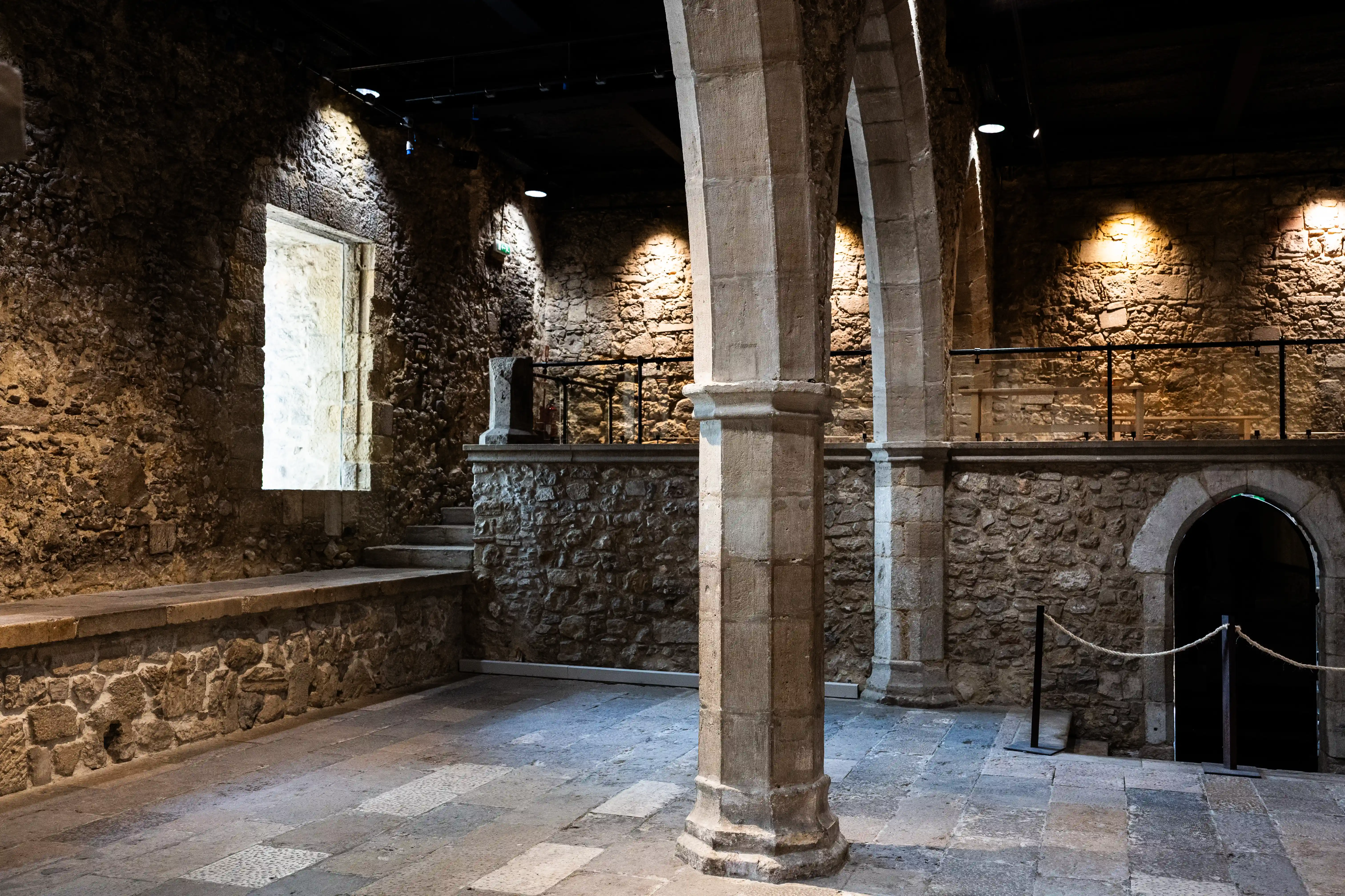  Interior view of a historic stone building with rough walls, large stone pillars, a narrow window, a staircase, and an arched doorway. Spotlights cast dramatic shadows.