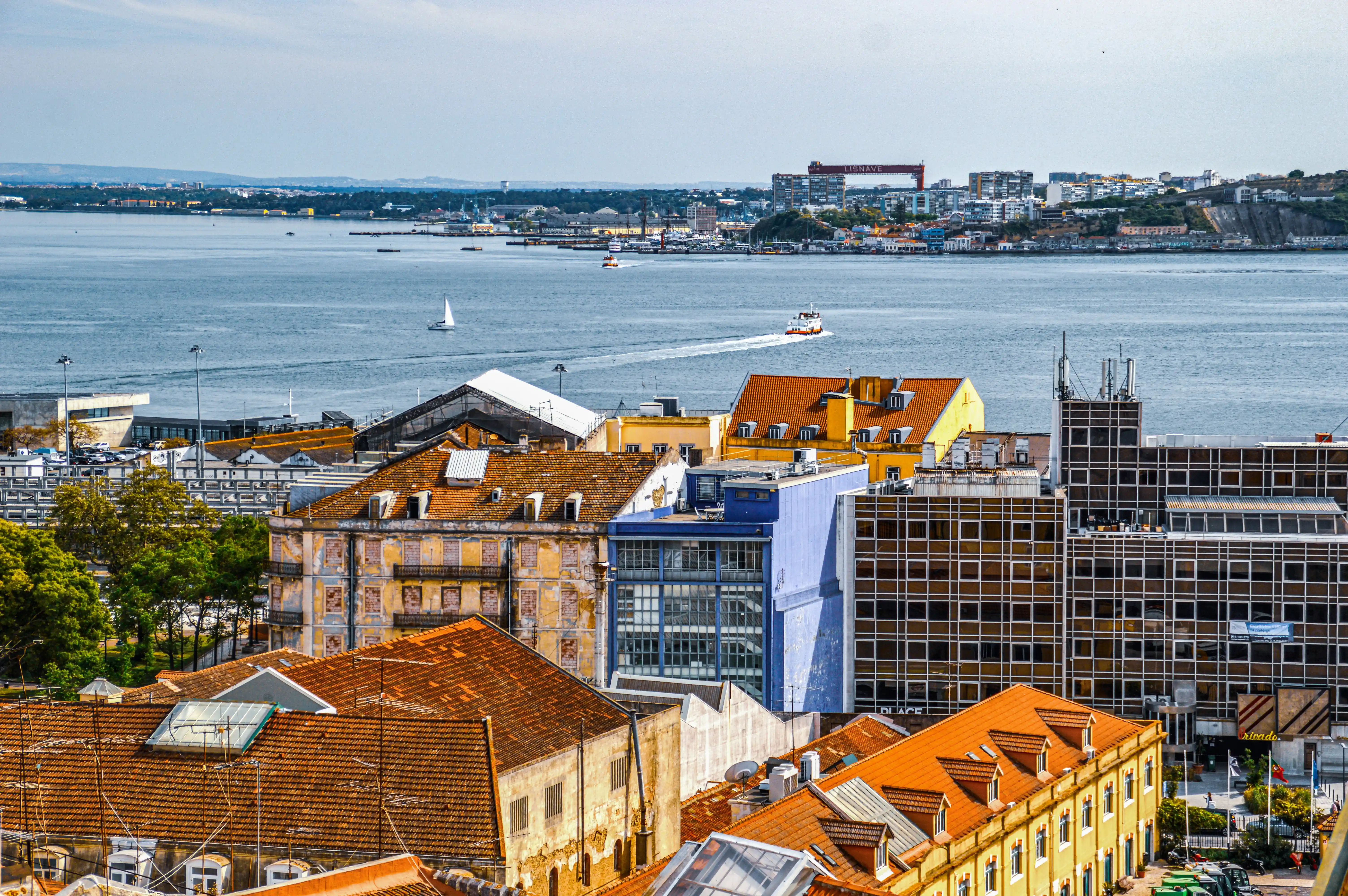 Aerial view of Lisbon harbour showing terracotta-roofed buildings in the foreground, modern structures, cranes, and the Tagus River with a sailboat and ferry.