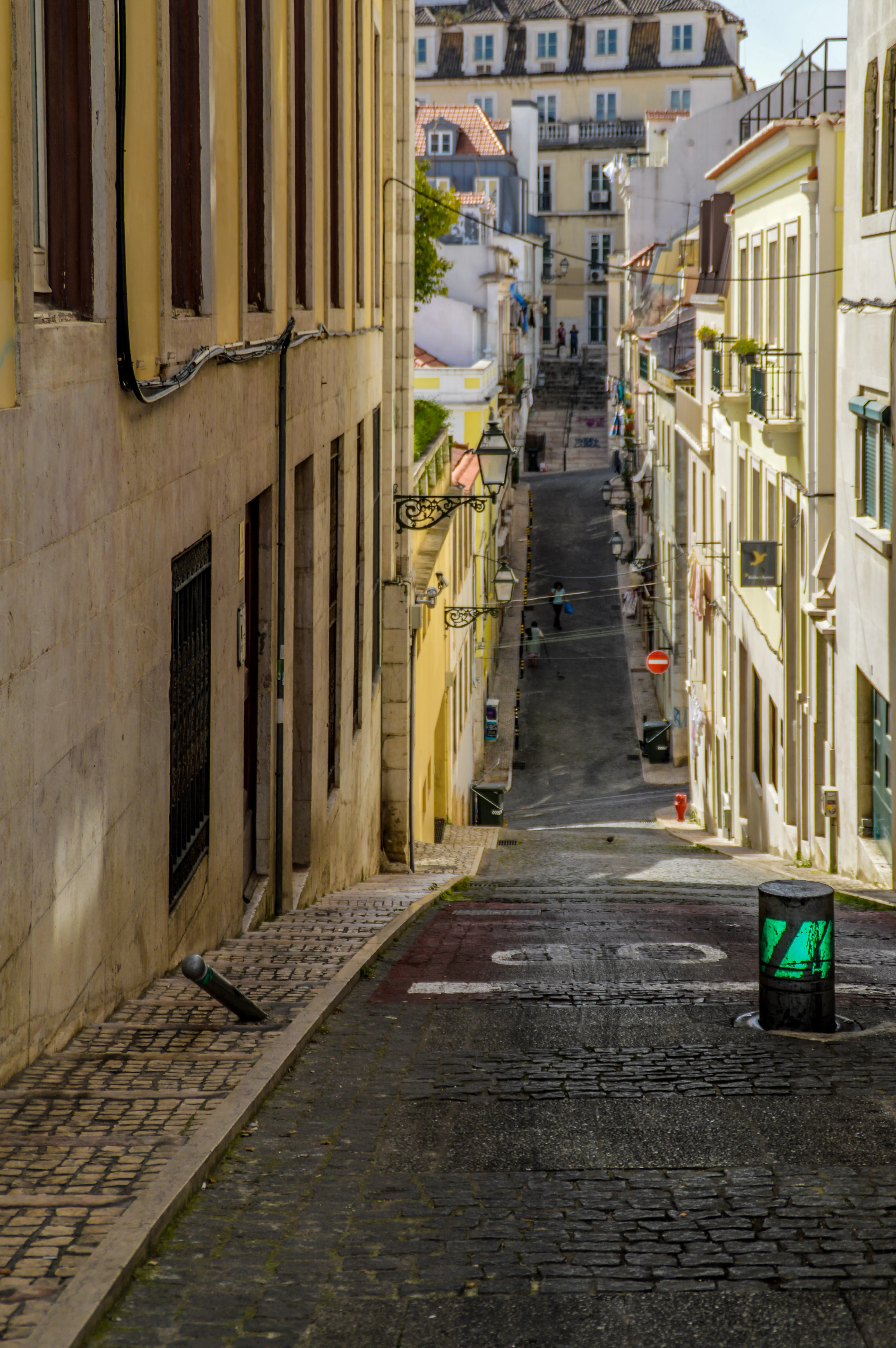 A steep, narrow cobblestone street lined with historic buildings, with pedestrians walking down the street. A green pedestrian traffic light is in the foreground.