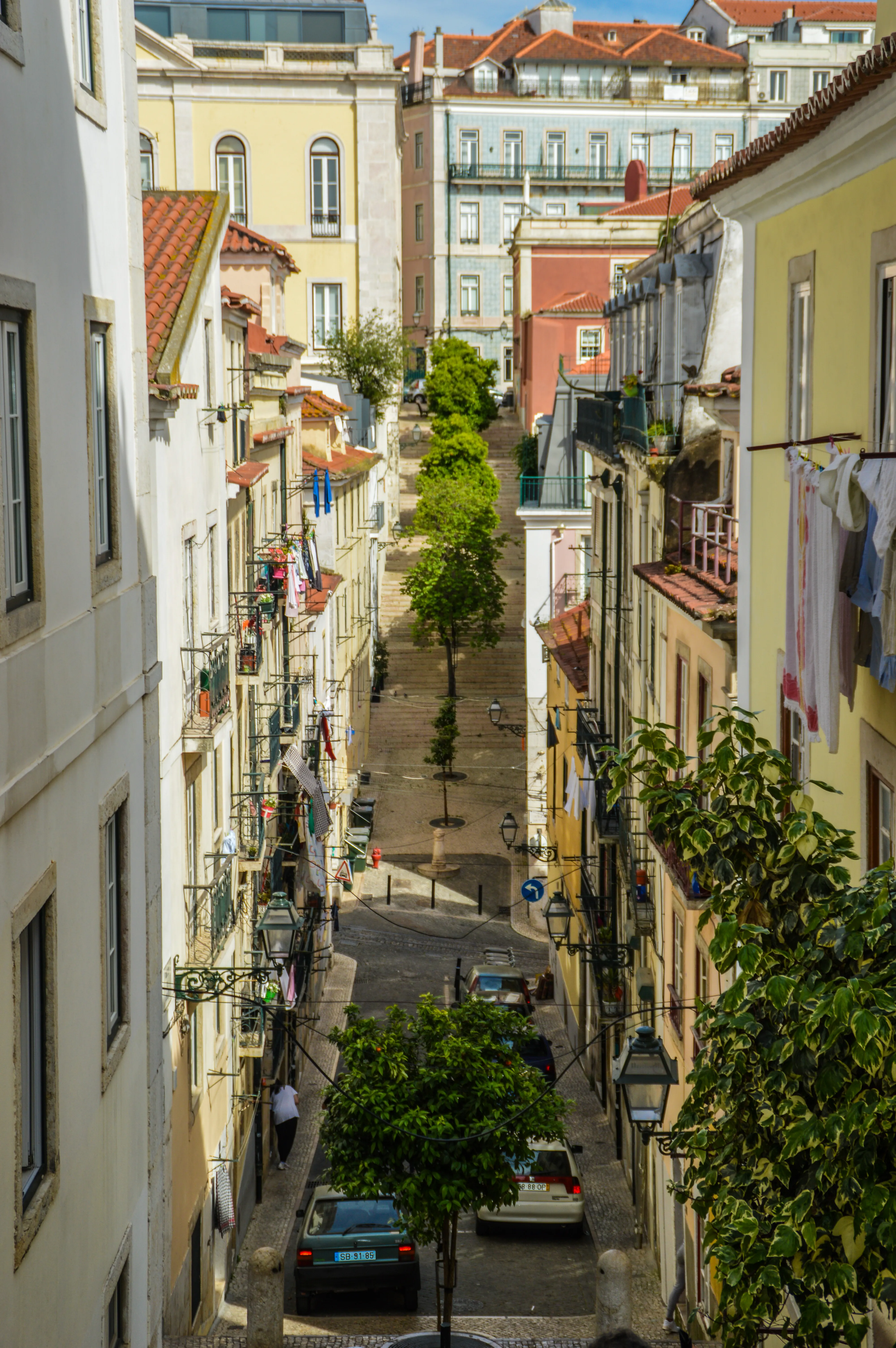 A view looking down a narrow, steep, cobblestone street in Lisbon, Portugal, with buildings of various pastel colours on either side. Laundry hangs from balconies, and trees are planted along the street. Cars are parked on the street.
