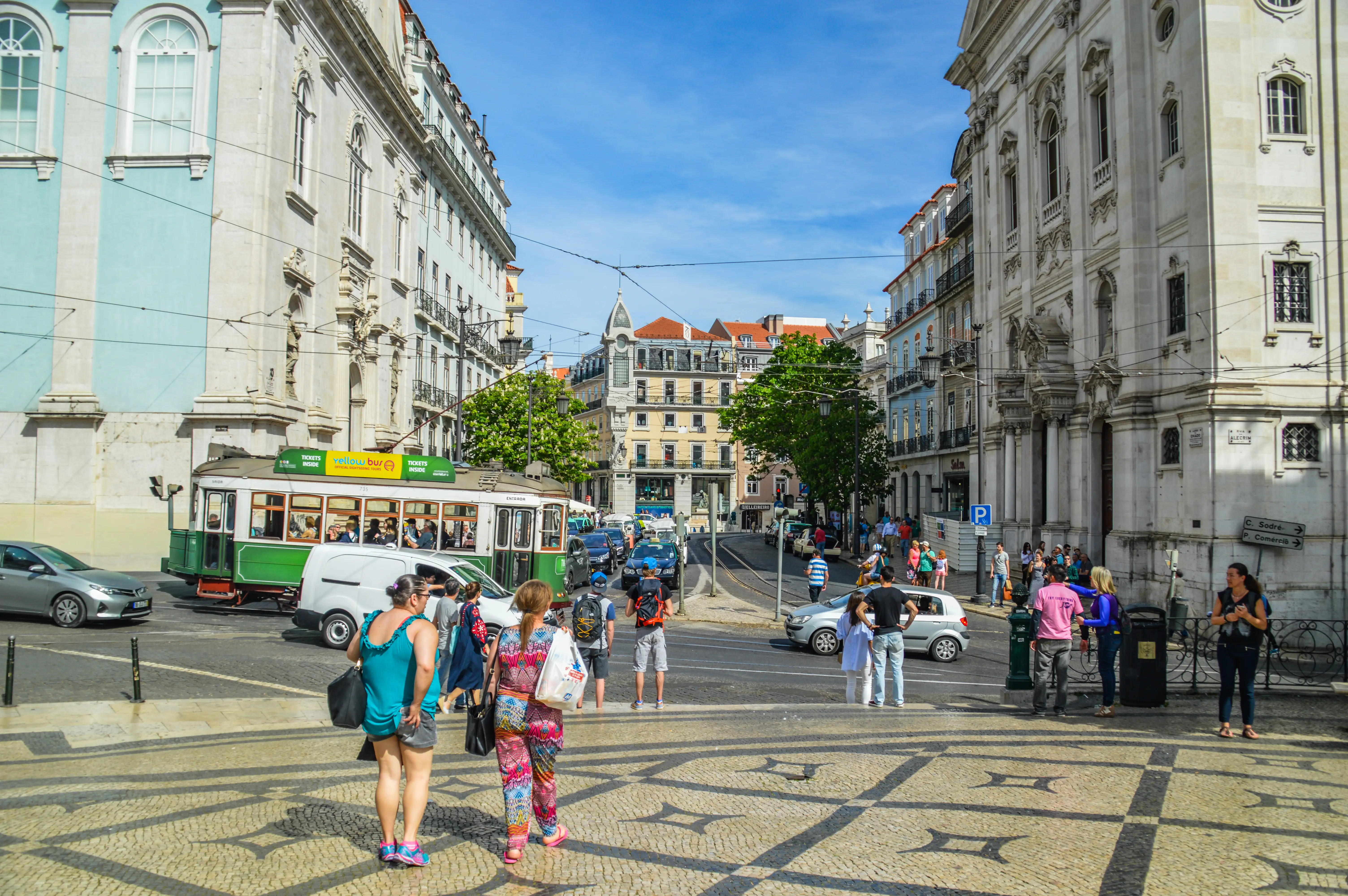 A wide shot of a sunlit street in Lisbon, Portugal, with a green and white tram and pedestrians on a mosaic pavement in the foreground, and grand, historical buildings lining both sides of the road.