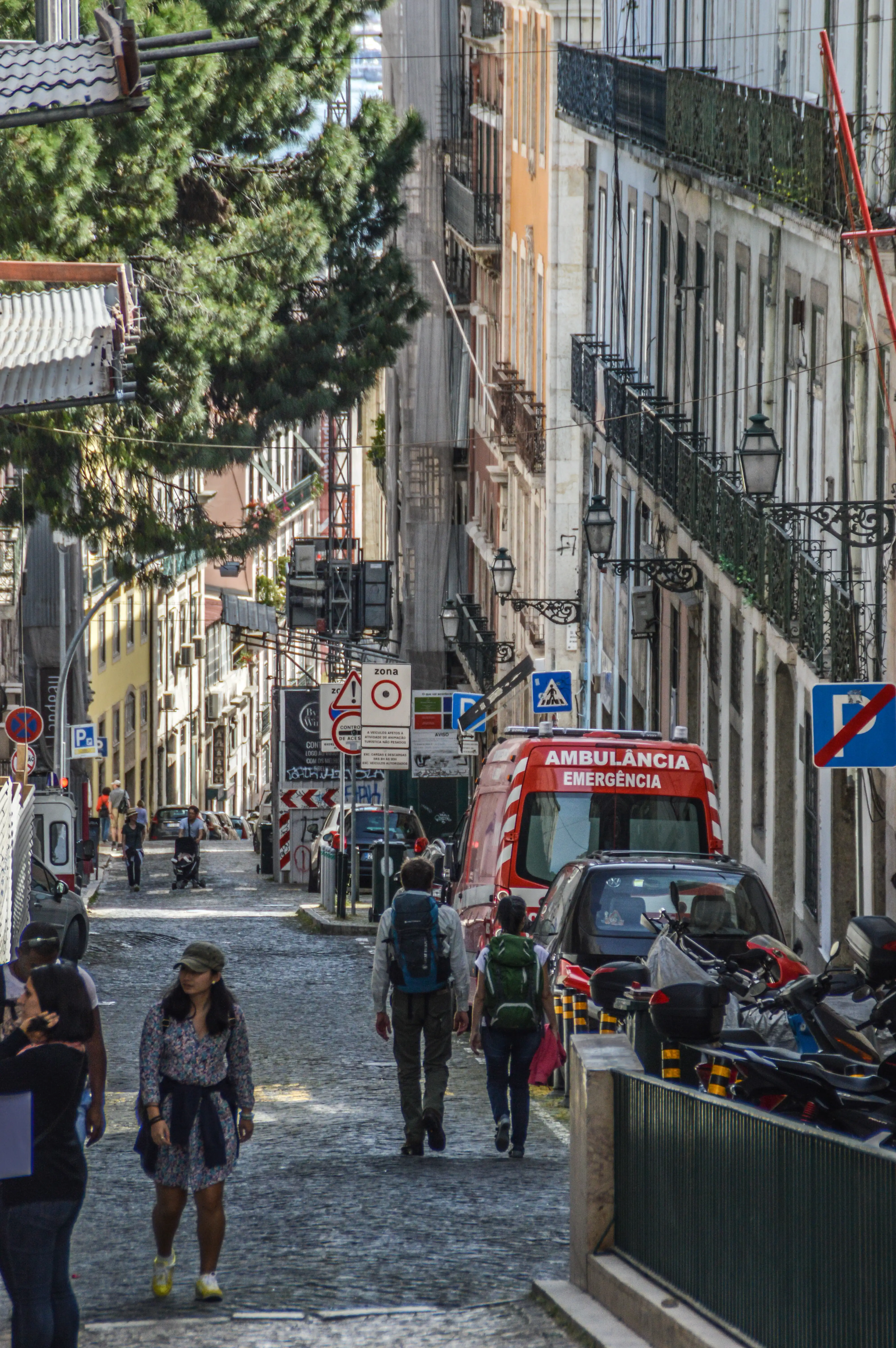A cobblestone street in Lisbon with buildings on either side, an ambulance, and several people walking.