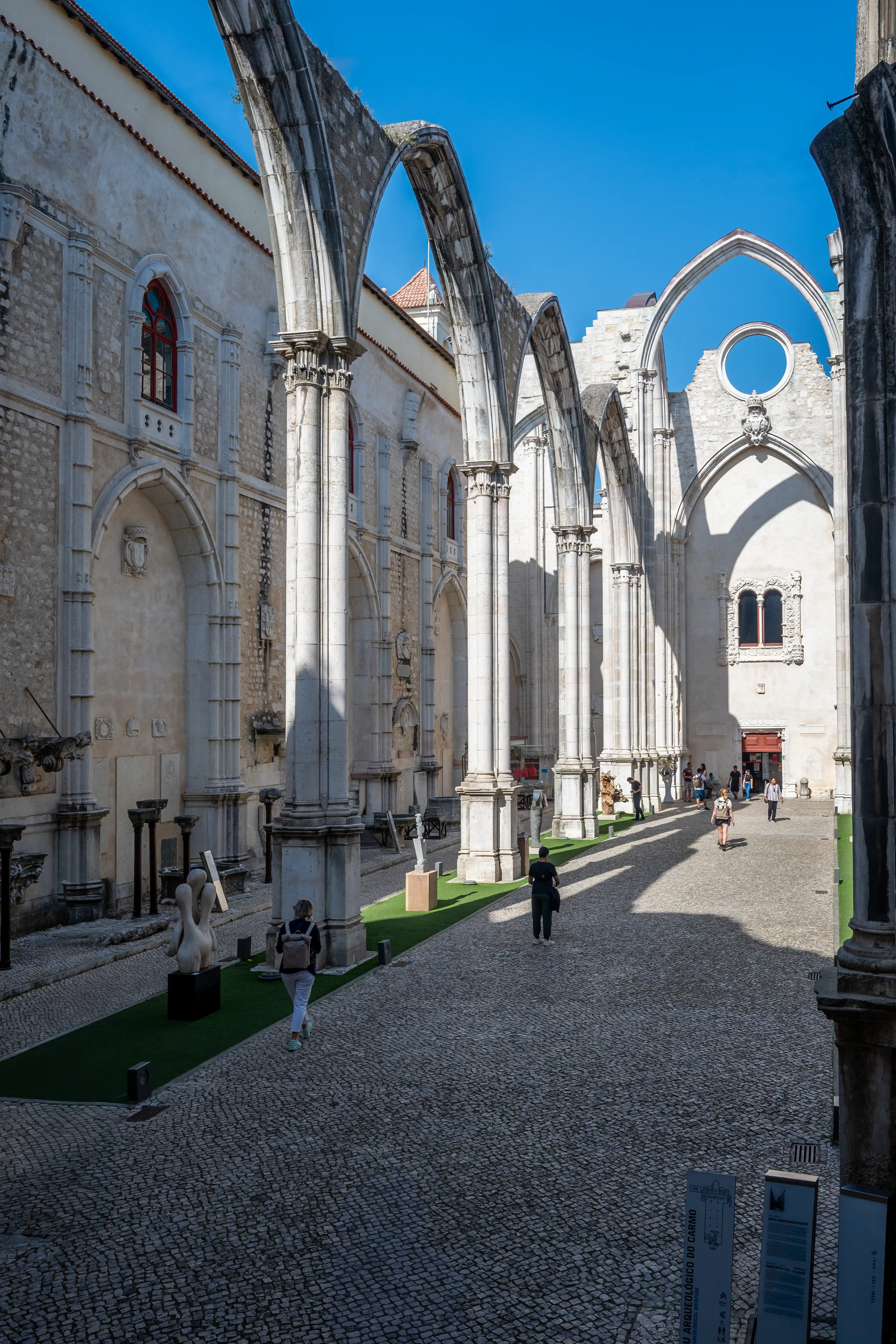 Exterior view of a ruined Gothic church courtyard with large stone arches, columns, cobblestone ground, green grass areas, sculptures, and a few people walking.