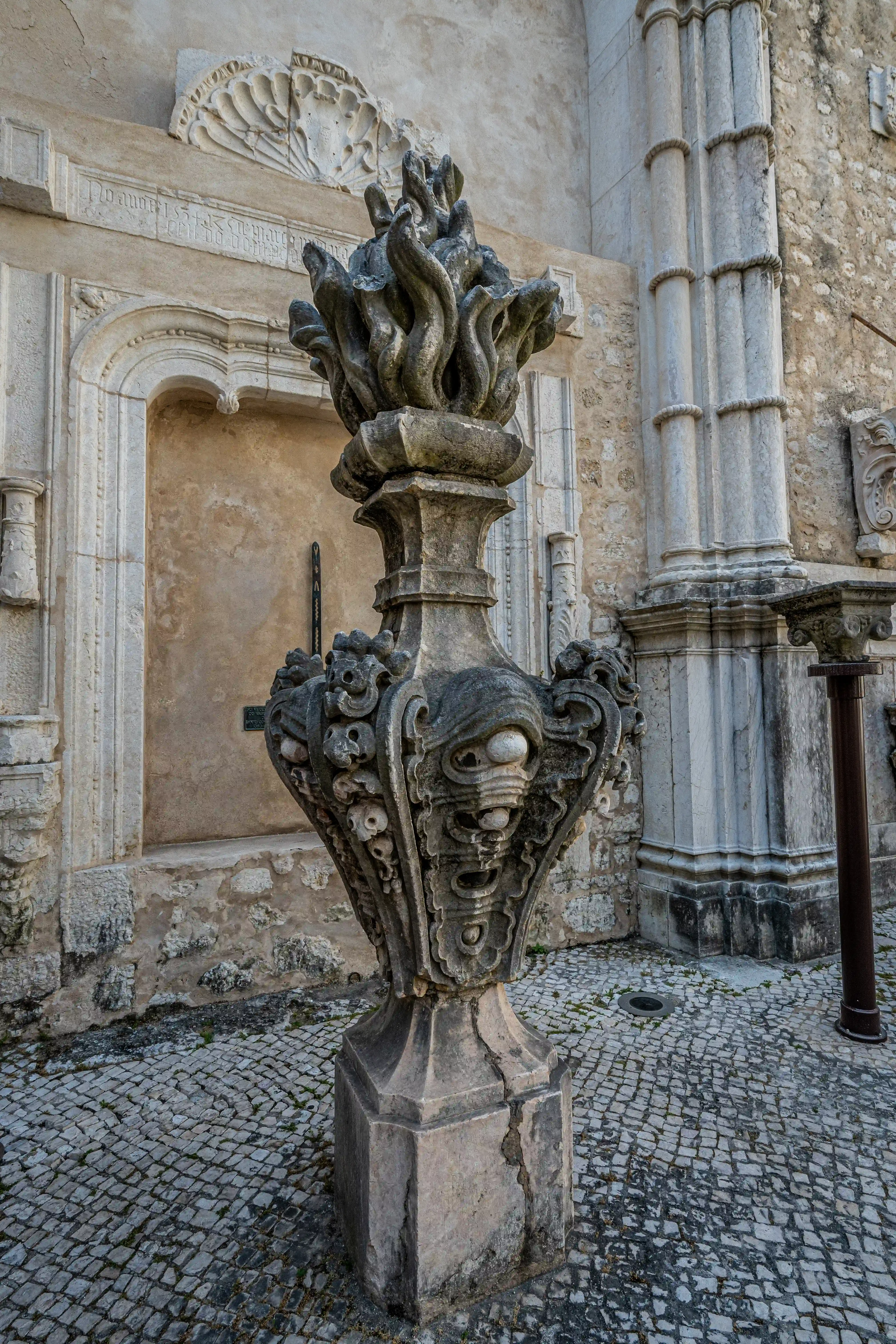 A weathered, ornate stone fountain with a sculpted flame motif at the top, set against a textured stone building wall and cobblestone pavement.