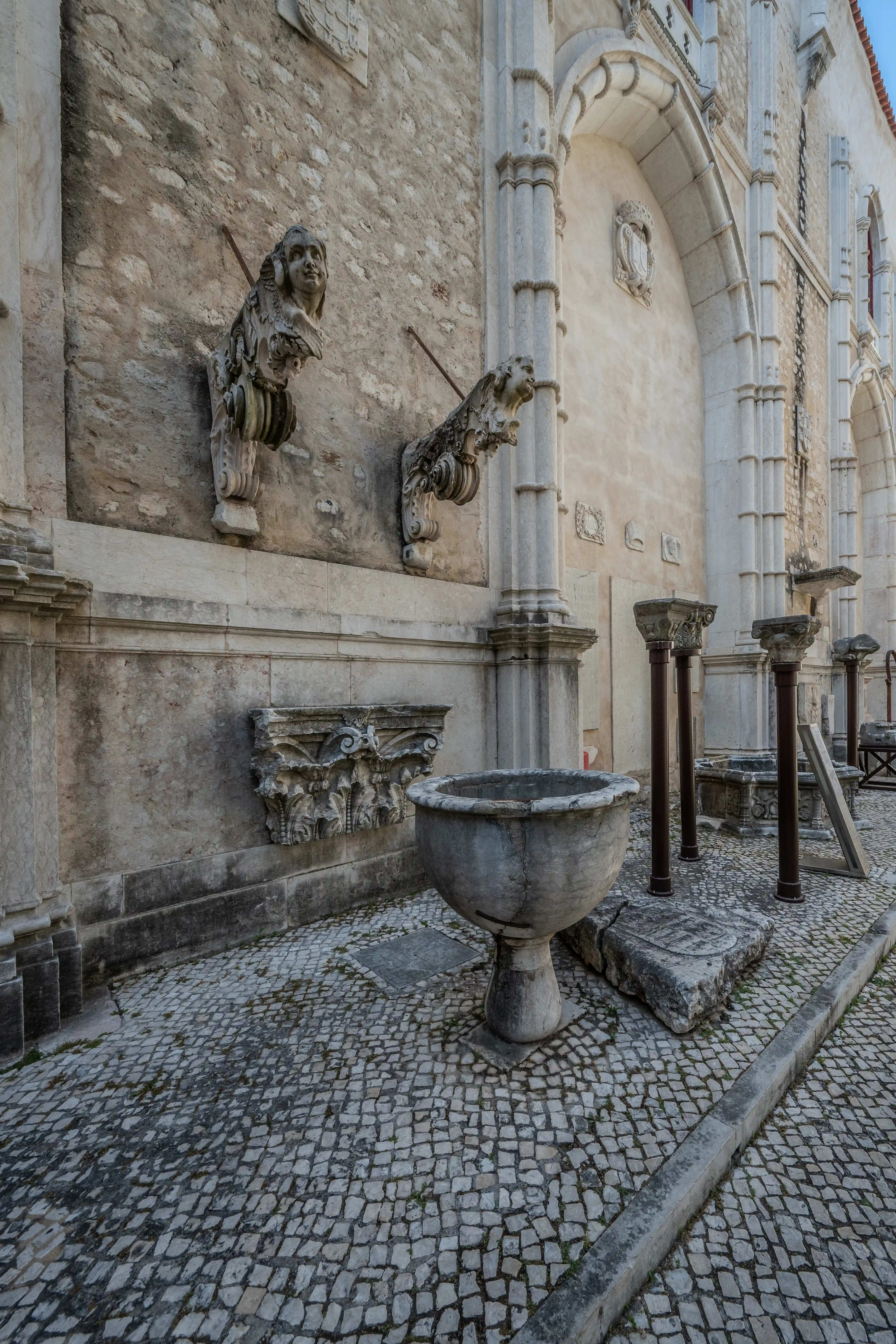 tone gargoyles with human and animal features mounted on a textured wall, a large stone fountain in the foreground, and columns and architectural remnants on a cobblestone pavement in front of an arched building.