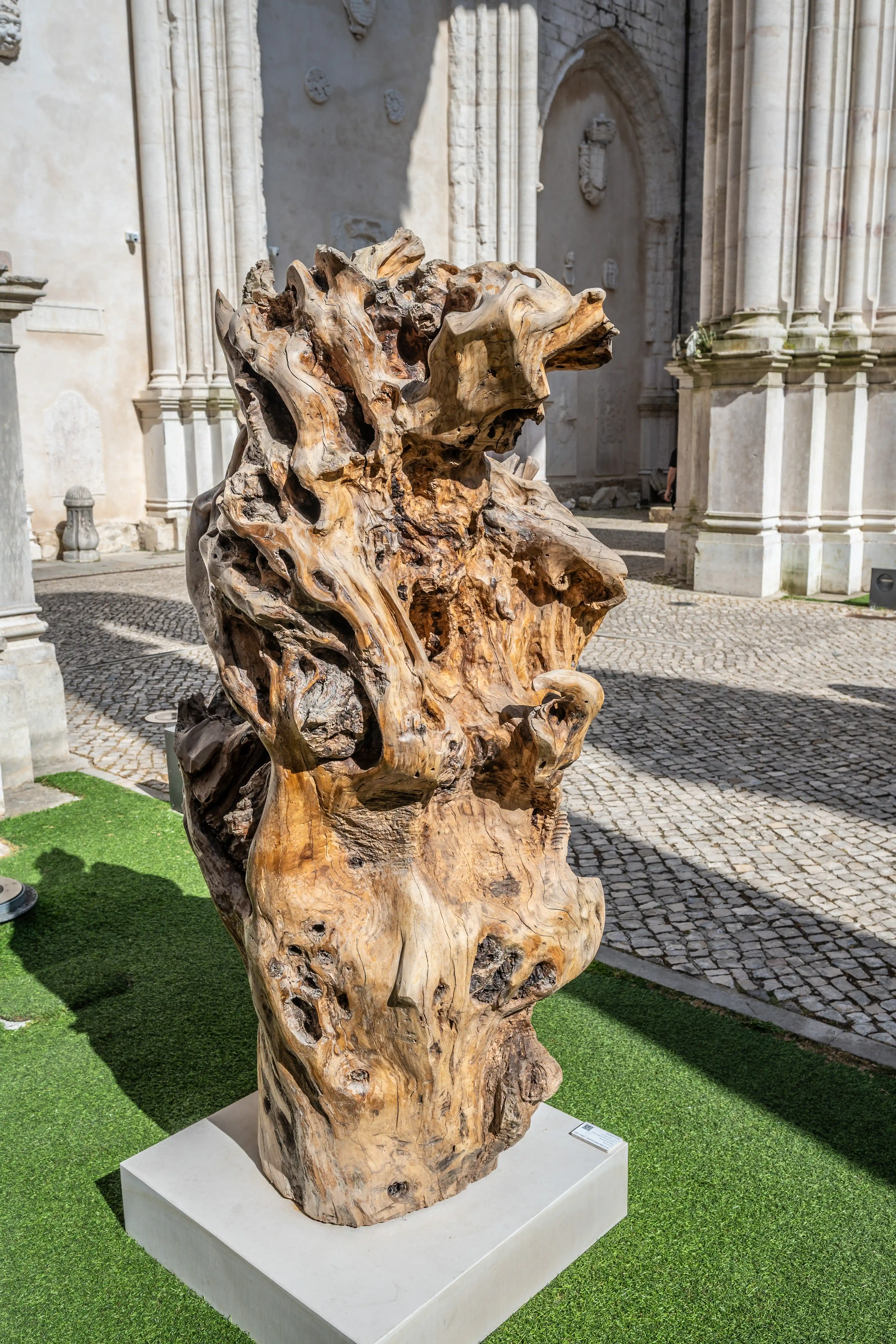 A large, complex sculpture made from a natural piece of wood, exhibiting a multitude of holes and twists, stands on a white plinth on artificial grass. A pale stone building with architectural details is in the background.