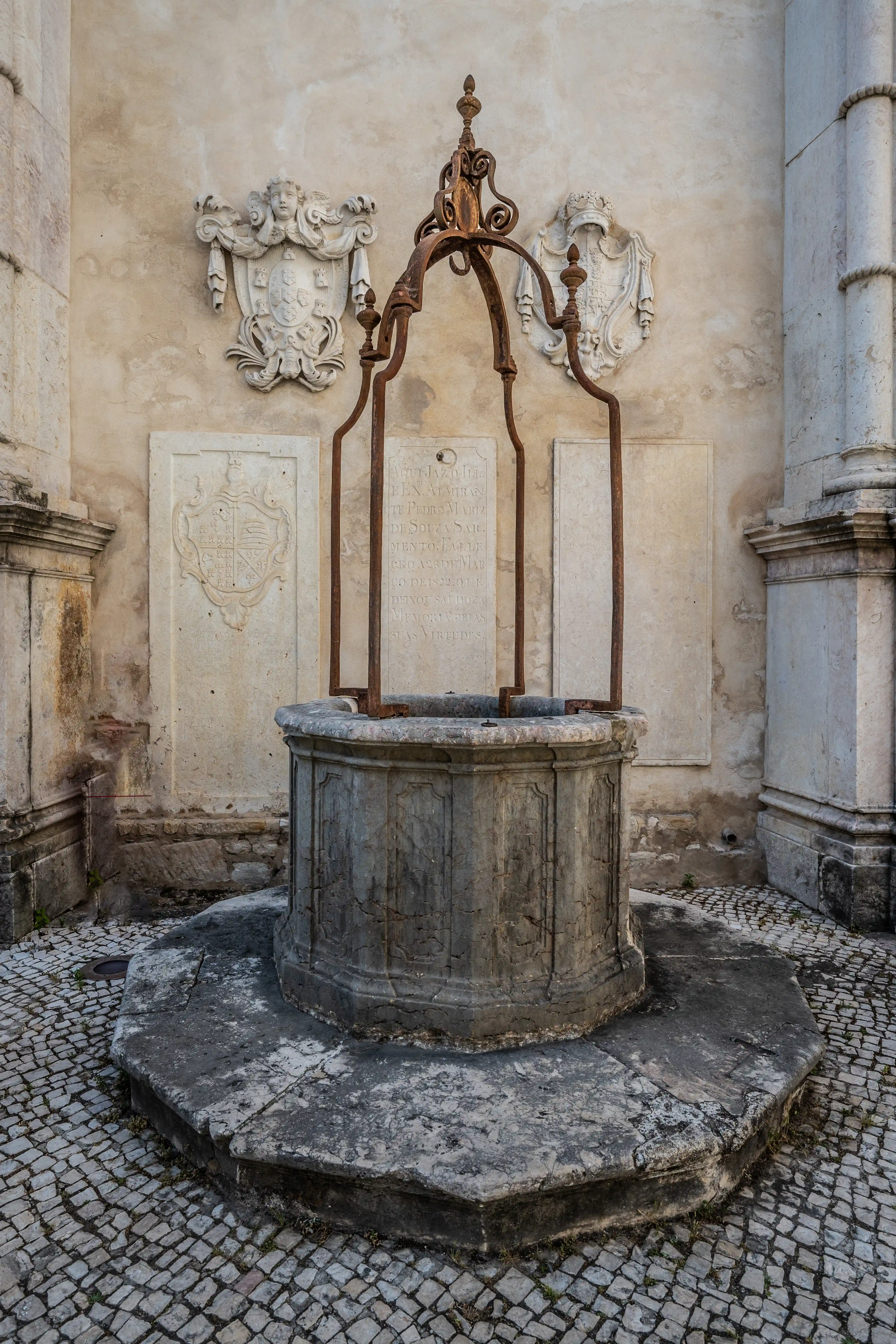 A stone well with an ornate rusty metal frame, set against a wall decorated with sculpted coats of arms and an inscription. Cobblestone paving surrounds the well.