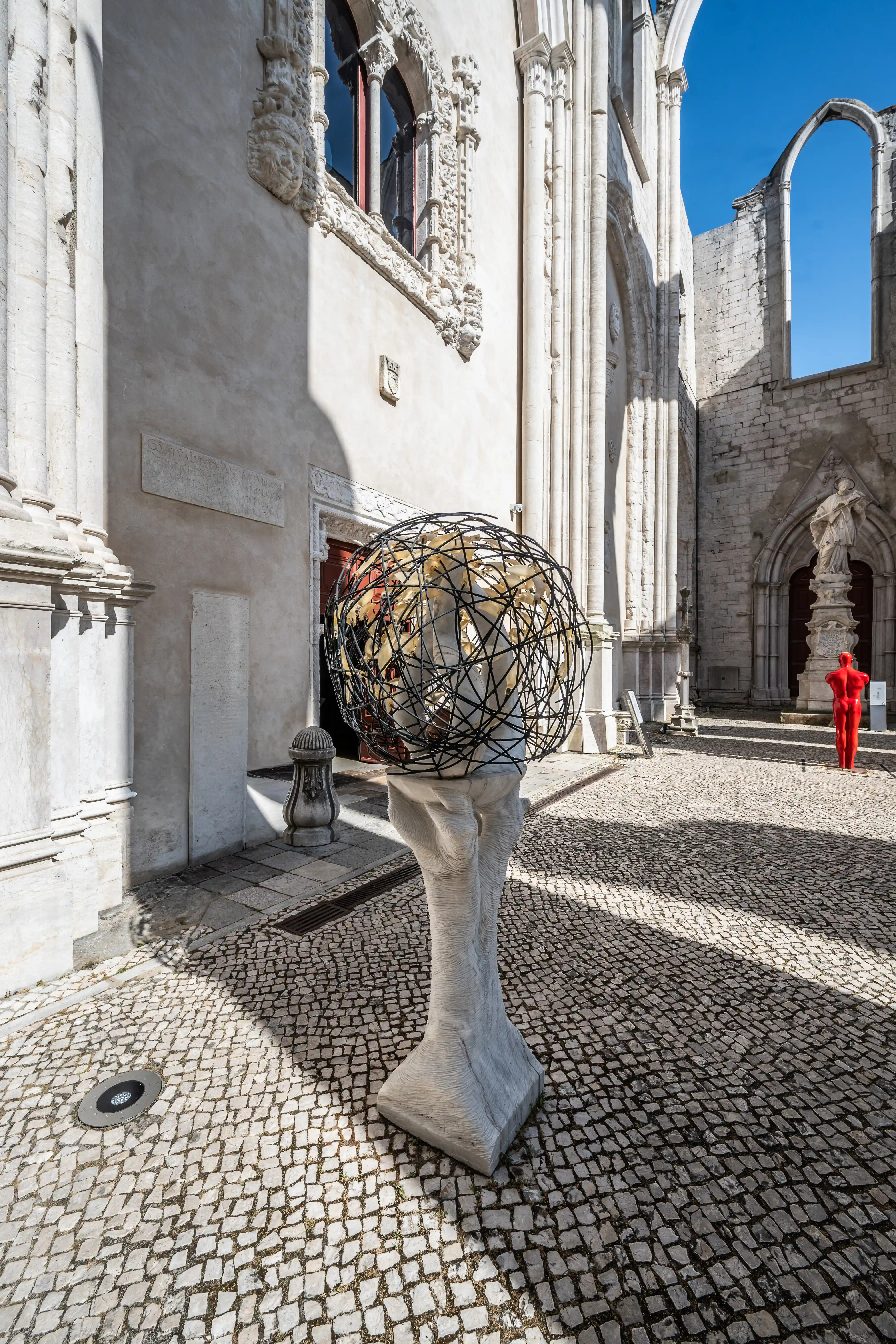 A contemporary sculpture, a sphere of black wire on a white pedestal, stands in the foreground of the partially ruined Gothic Carmo Convent in Lisbon. Two people sit on a bench to the left, and a bright red human figure stands to the right.
