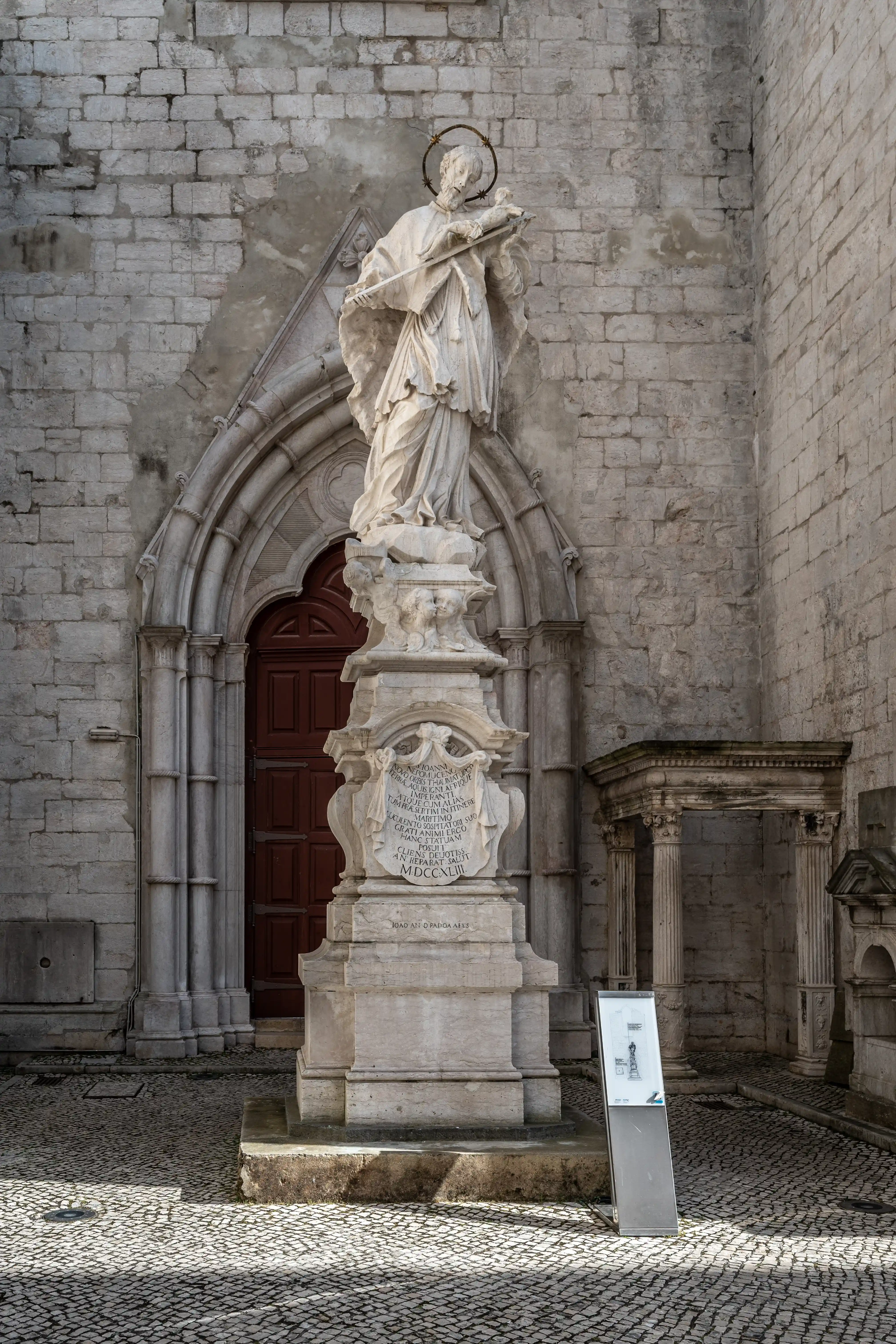 A white stone statue of a saint with a halo stands on a pedestal in front of a large arched wooden door and a stone building. Paved cobblestone ground.