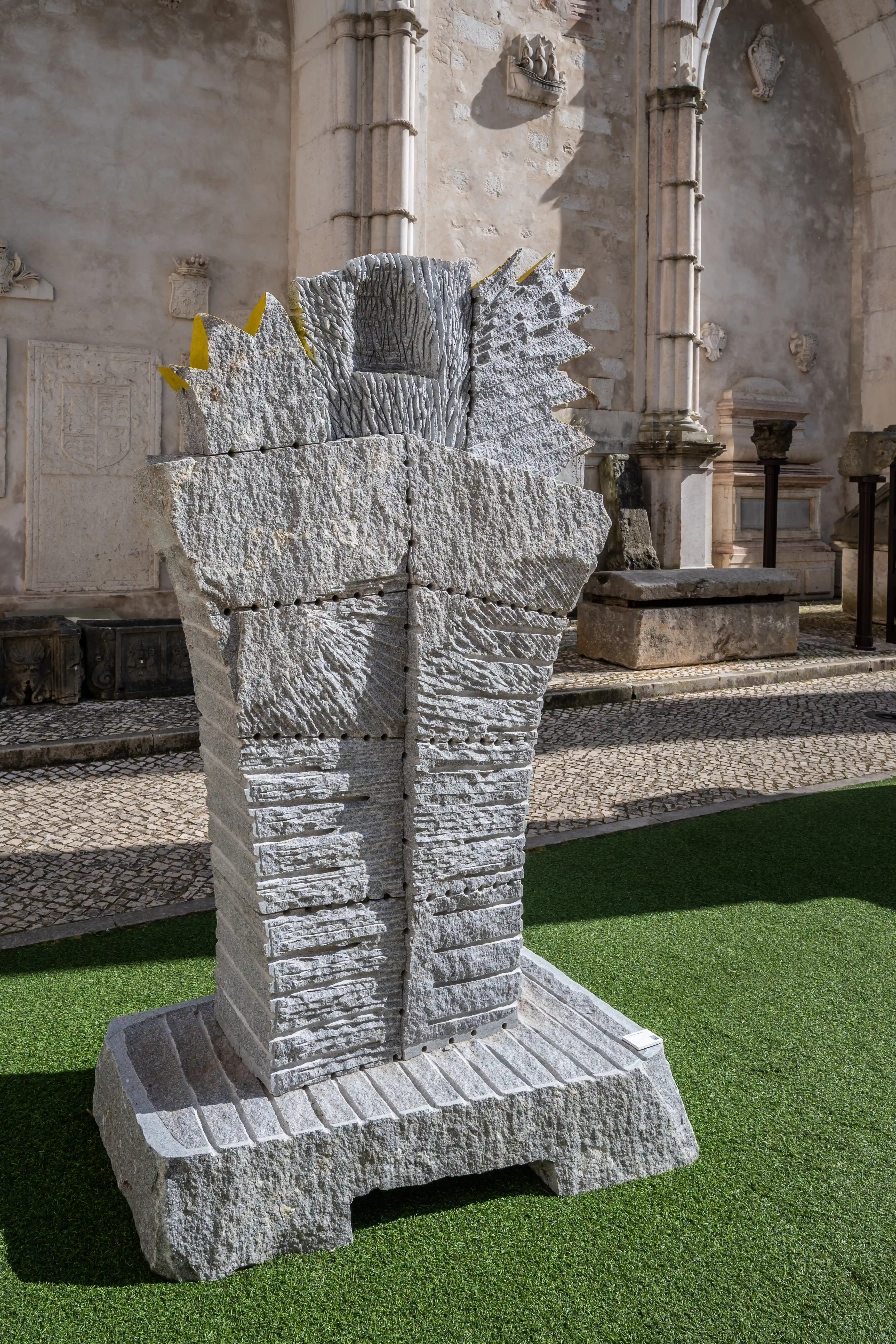 An abstract grey granite sculpture with a rough, carved texture is placed on artificial grass in front of an old stone building with architectural features.