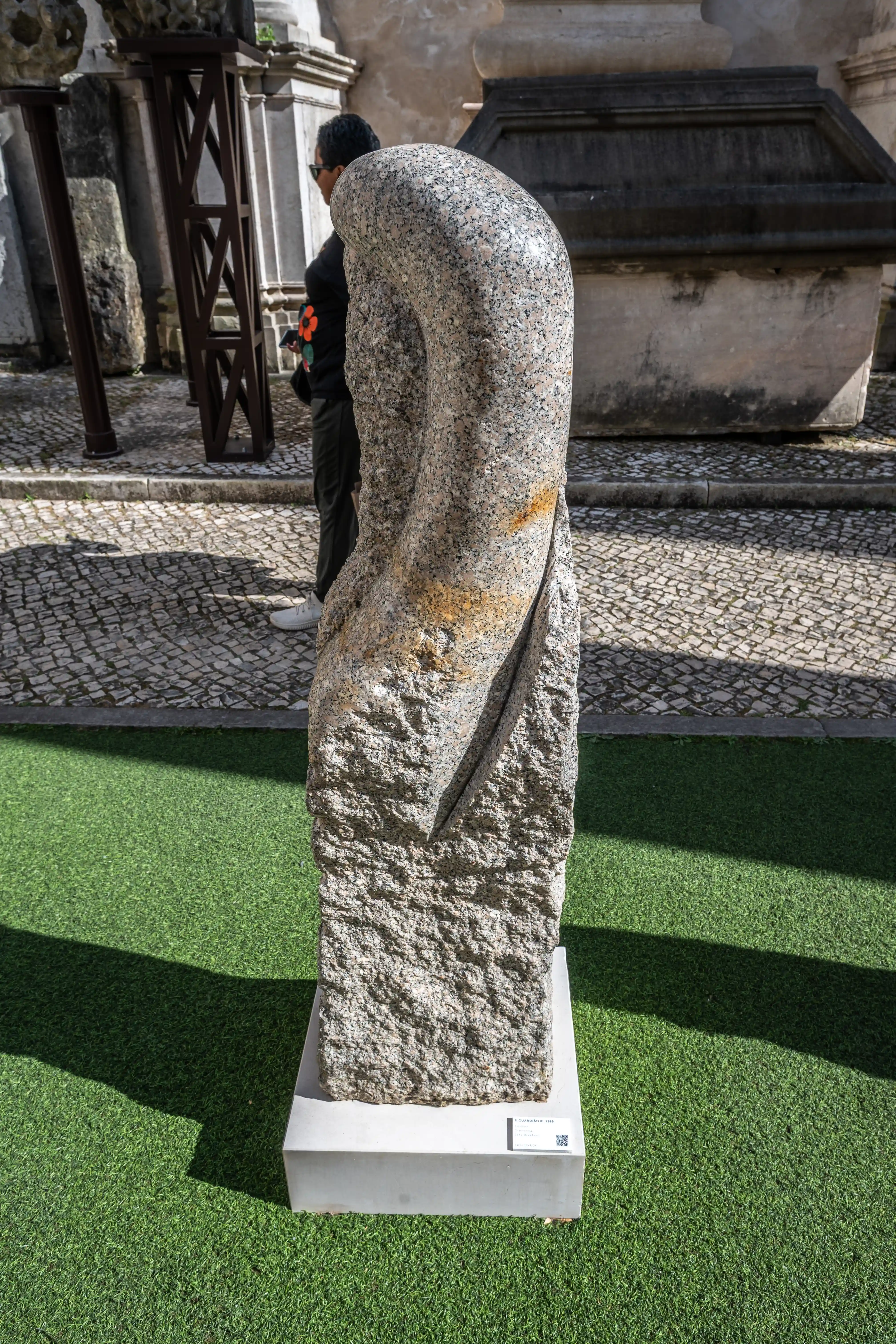 A grey granite abstract sculpture with a smooth, curved top section and a rough, textured base, displayed outdoors on a green artificial turf surface.