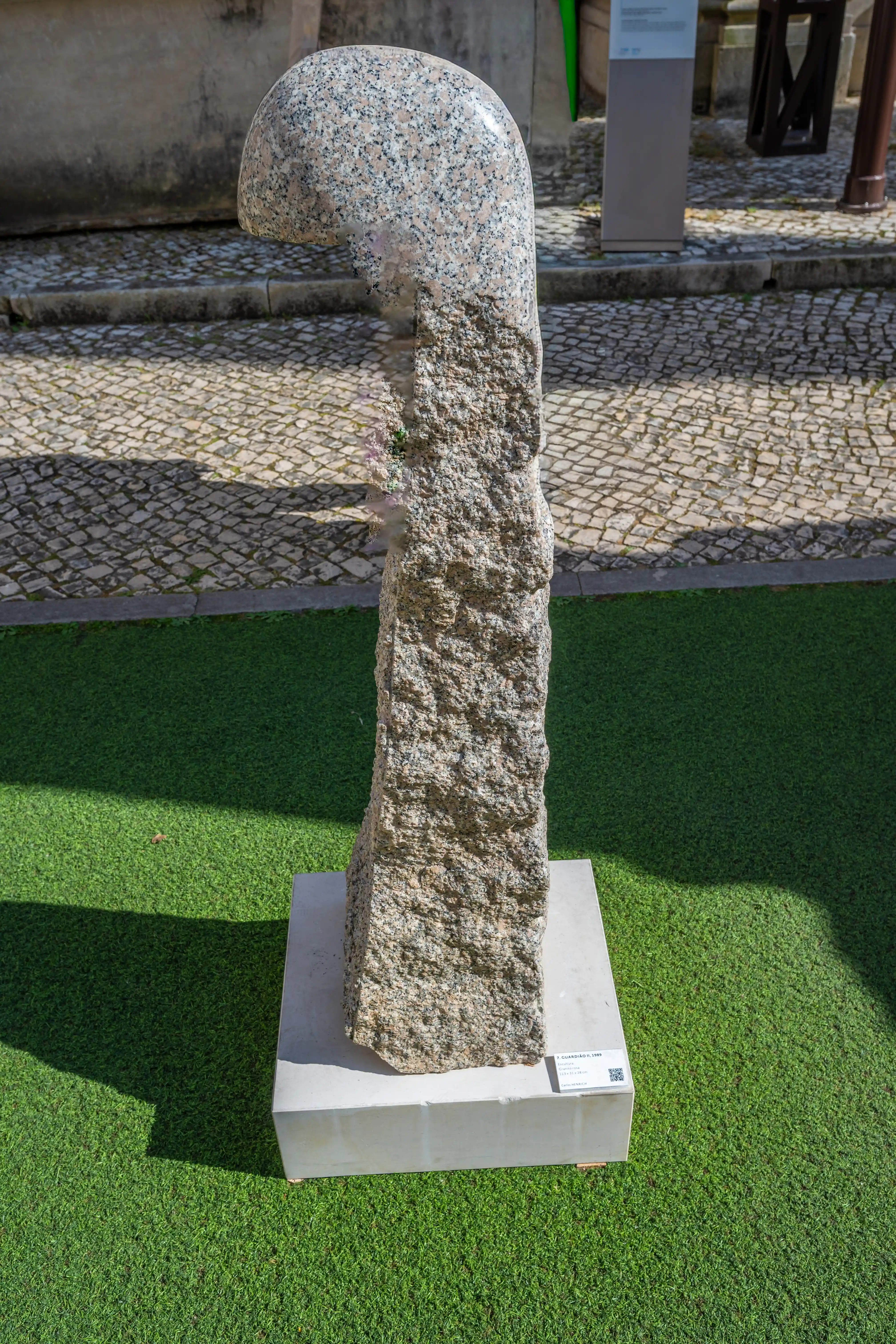 A grey granite abstract sculpture with a smooth, curved top section and a rough, textured base, displayed outdoors on a green artificial turf surface.