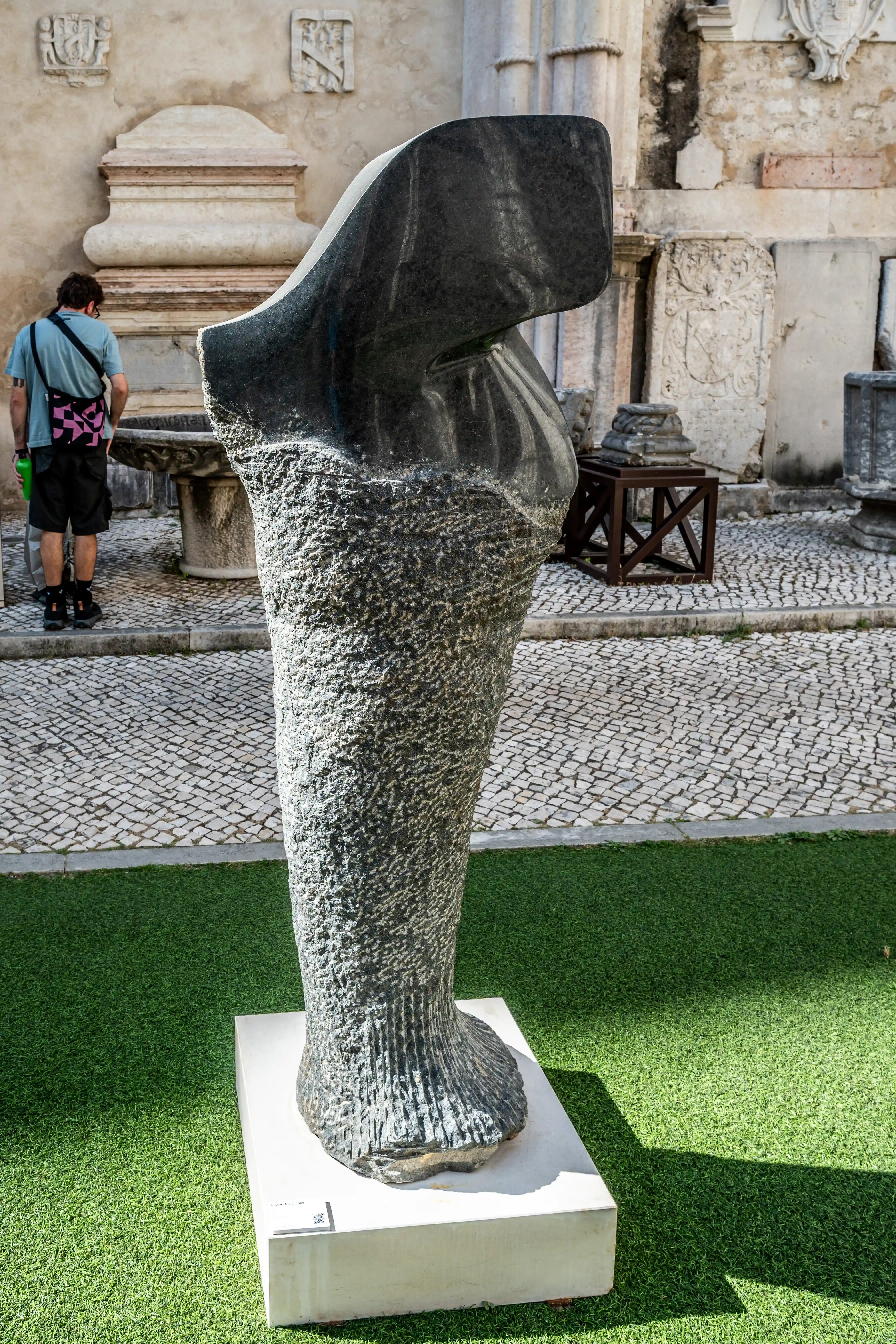 A dark, abstract stone sculpture with a rough texture is displayed outdoors on a white pedestal. In the background, a person stands near a stone fountain in a courtyard paved with cobblestones.