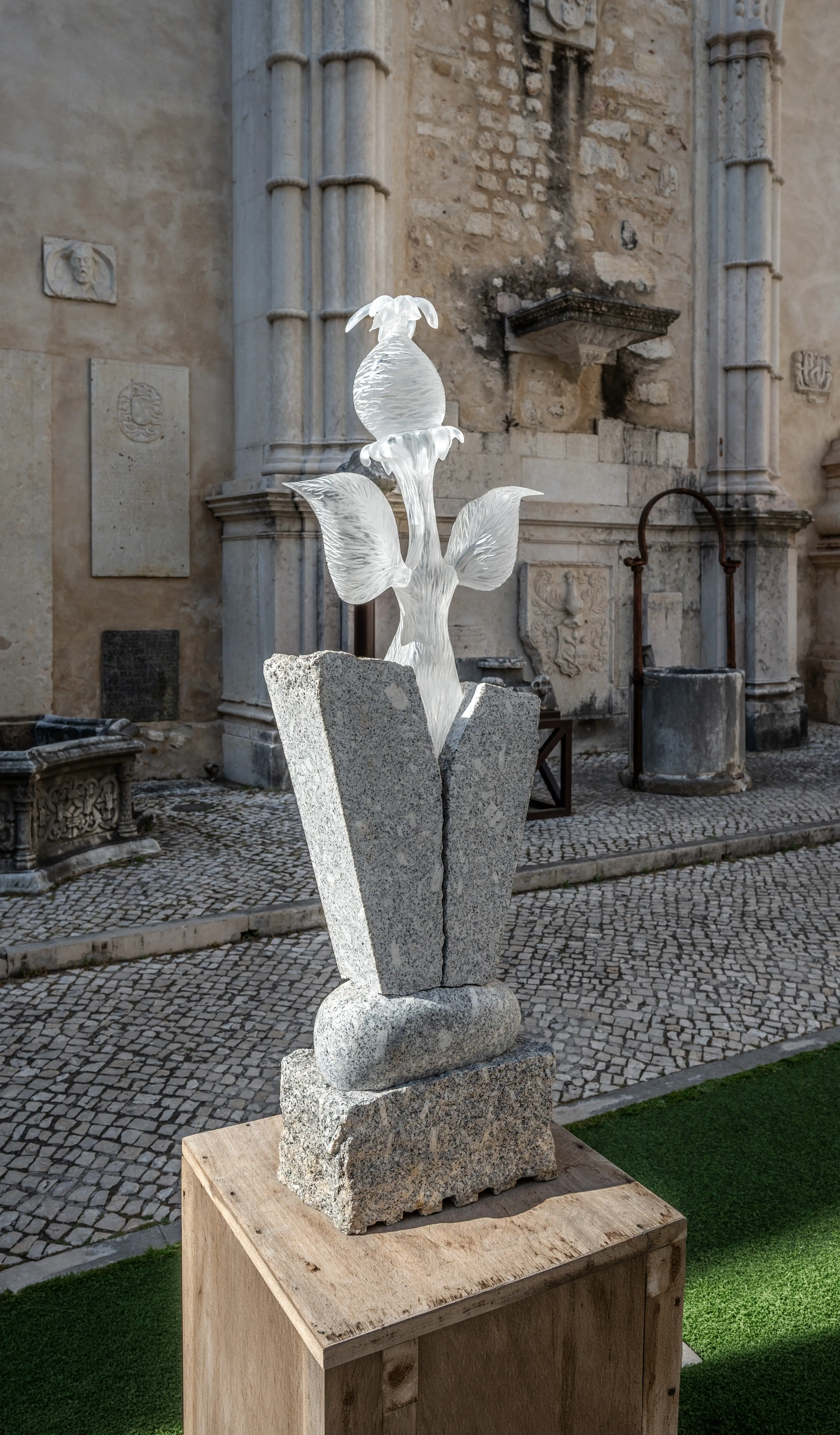 A clear glass sculpture of a flower with textured petals and a bud-like top, mounted on a segmented grey granite base. The sculpture is set against a backdrop of an old stone building with architectural details and a cobblestone courtyard.