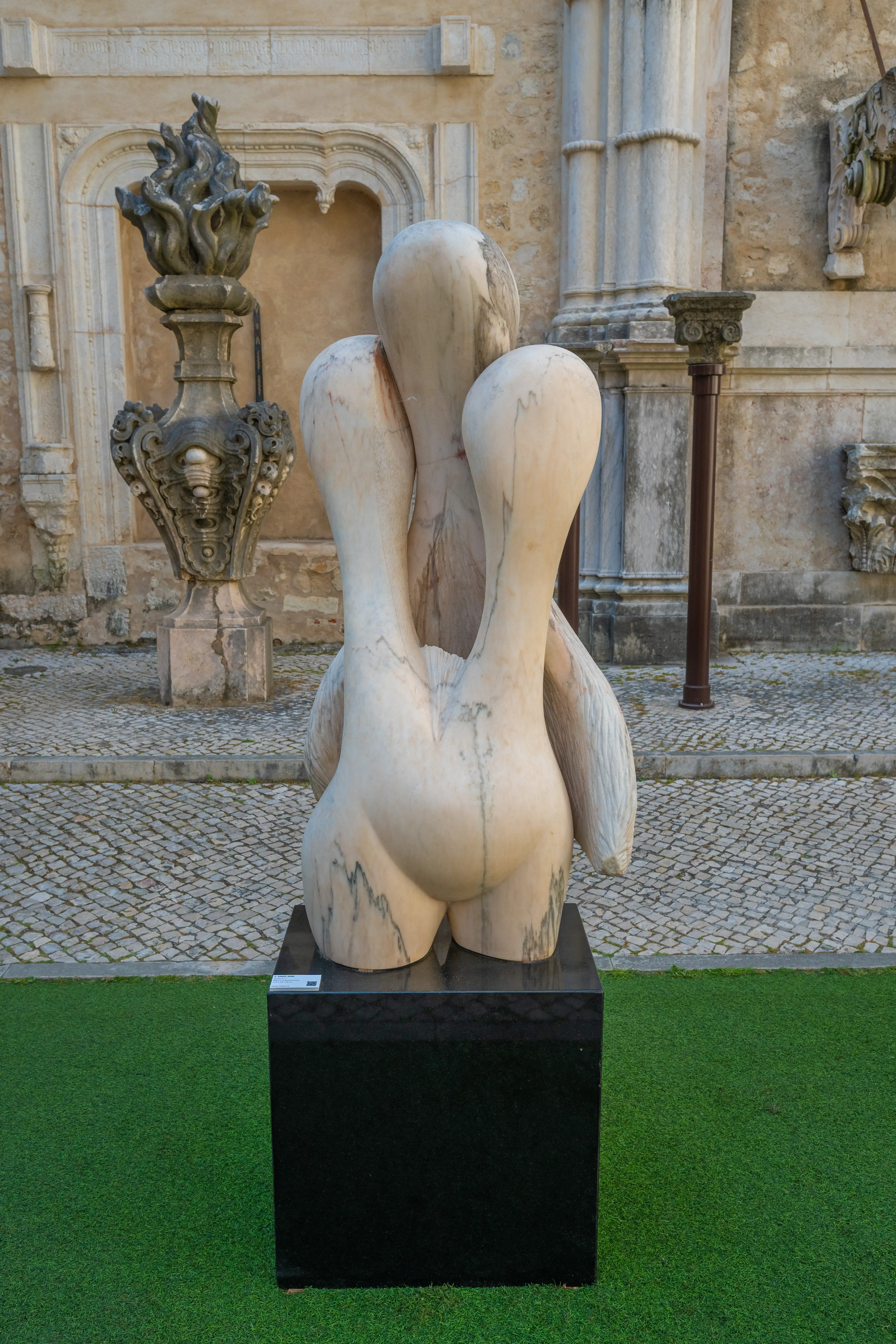 A view from behind of a white marble sculpture of three intertwined abstract figures, standing on a black plinth. To the left, a decorative stone fountain with sculpted flames. The background shows textured stone walls and columns of an old building, with cobblestone paving
