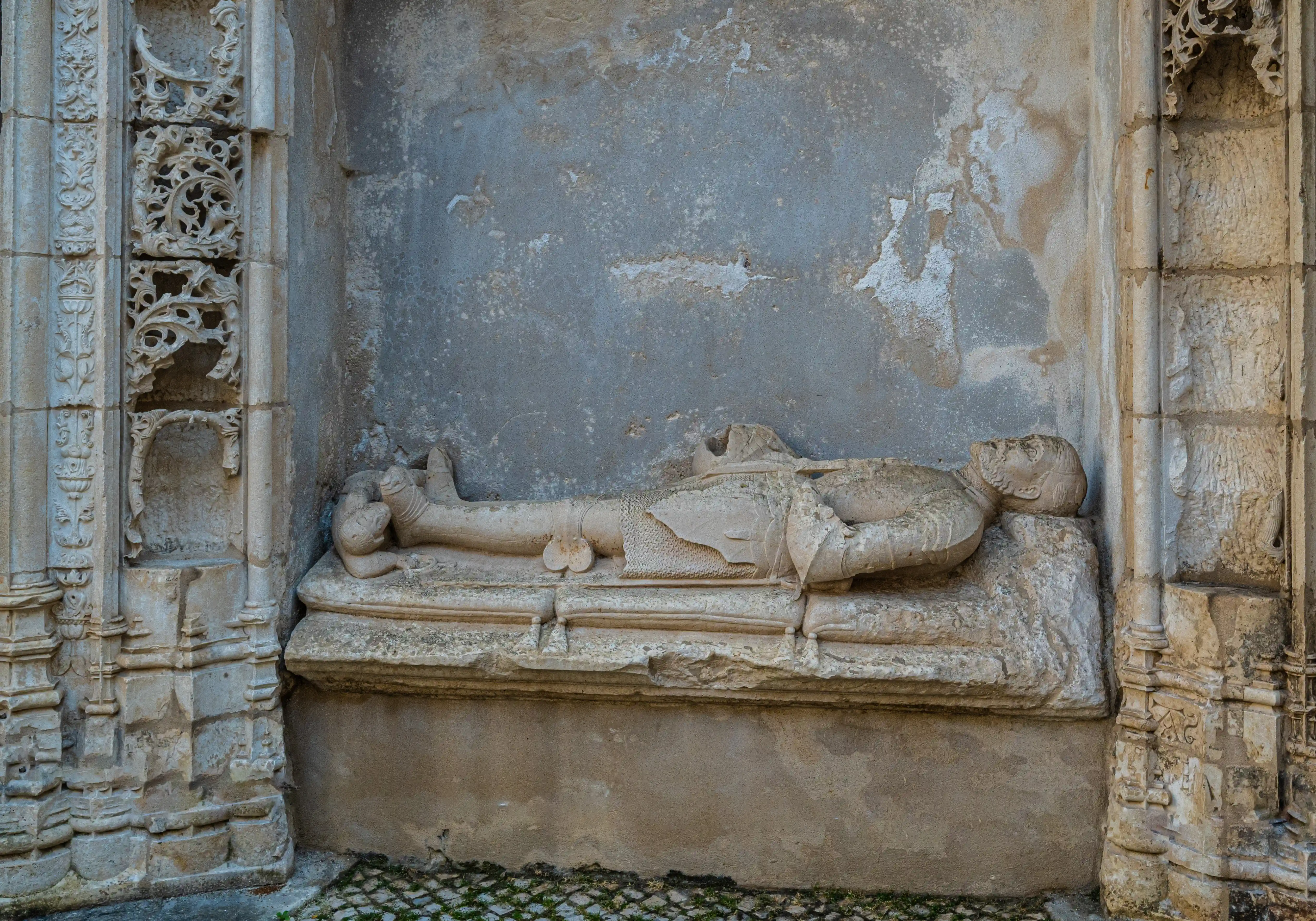  A stone tomb effigy of a knight in full armour, lying in a recessed architectural niche with detailed stonework, set against a textured wall, with a cobbled ground in the foreground.