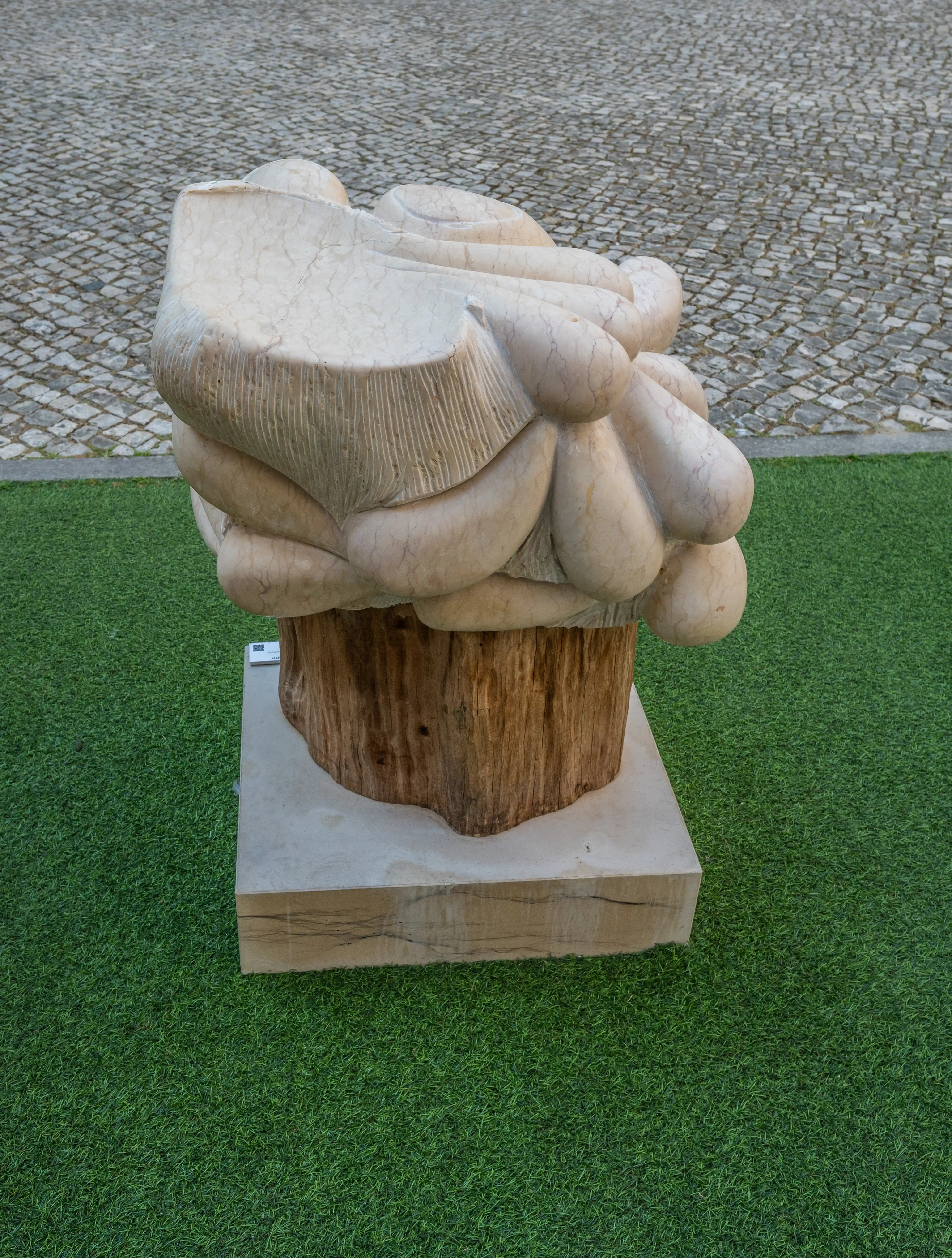 Abstract sculpture of pale marble with rounded shapes and carved lines, mounted on a wooden base, placed on green artificial turf.