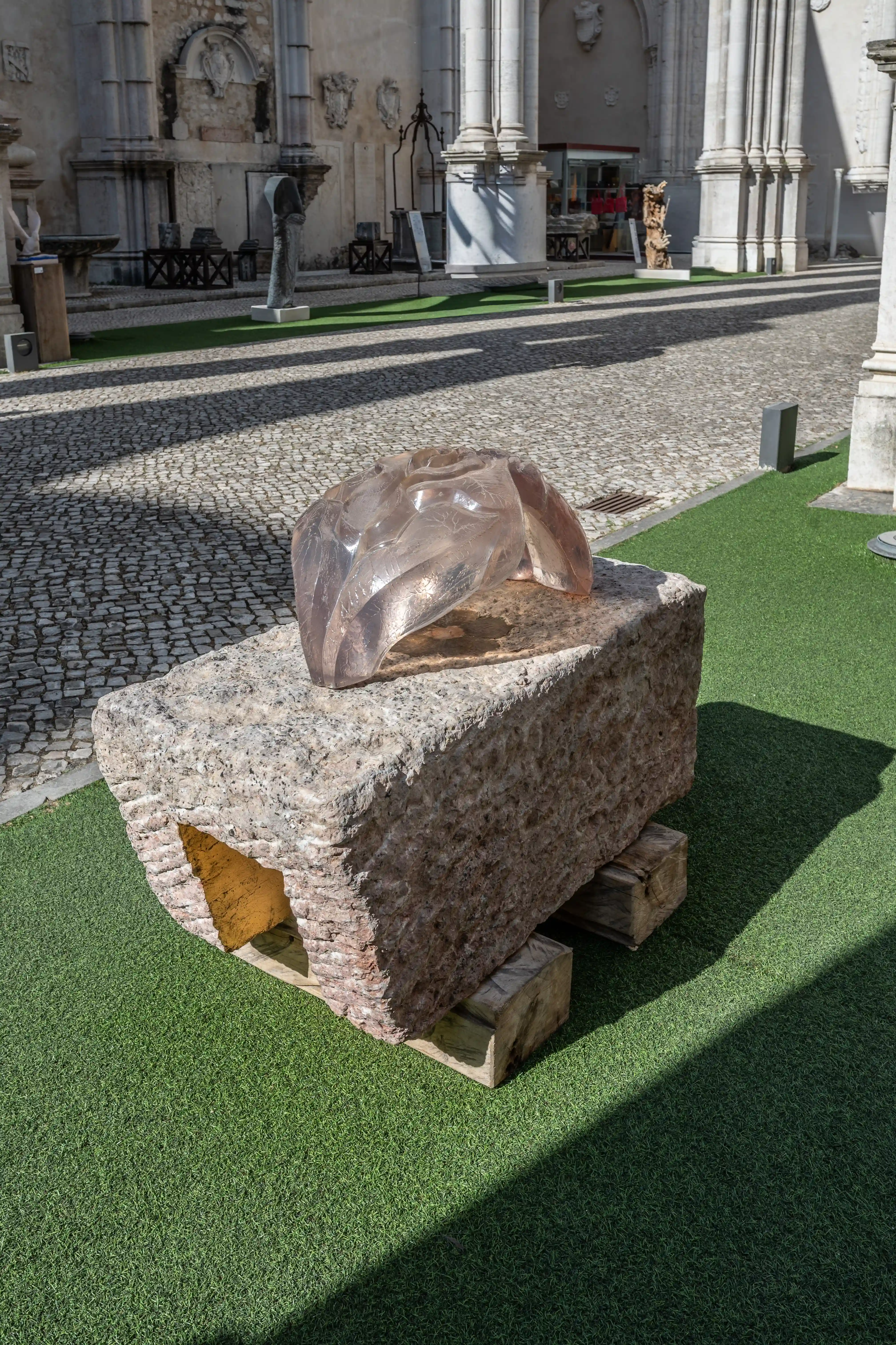 A contemporary sculpture made of translucent material rests on a large, coarse stone block in an open-air courtyard. People are visible in the background, observing the art.