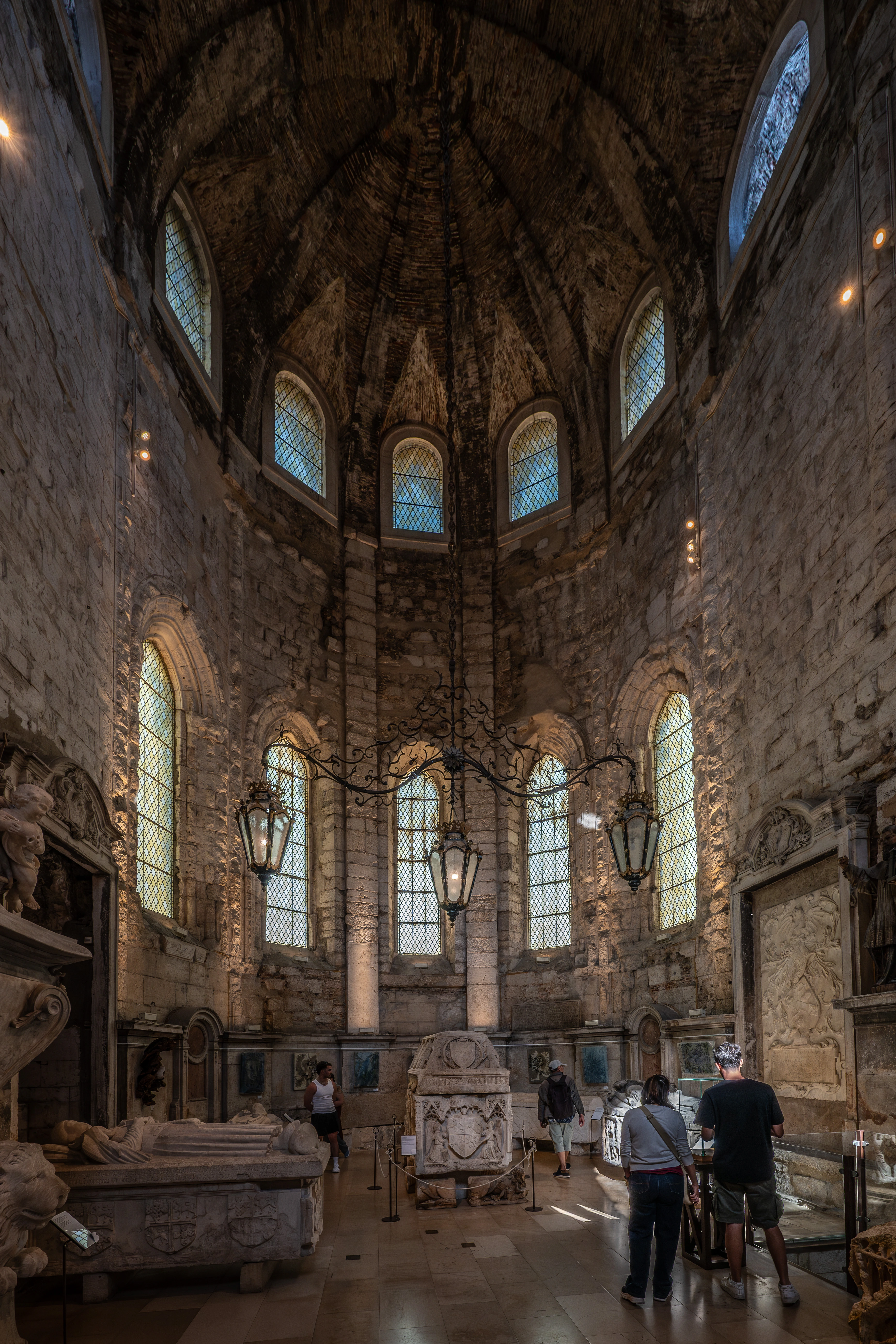 Interior view of a stone building with arched windows, ancient tombs, and a few visitors looking at the exhibits. Sunlight creates a dramatic play of light and shadow on the floor.