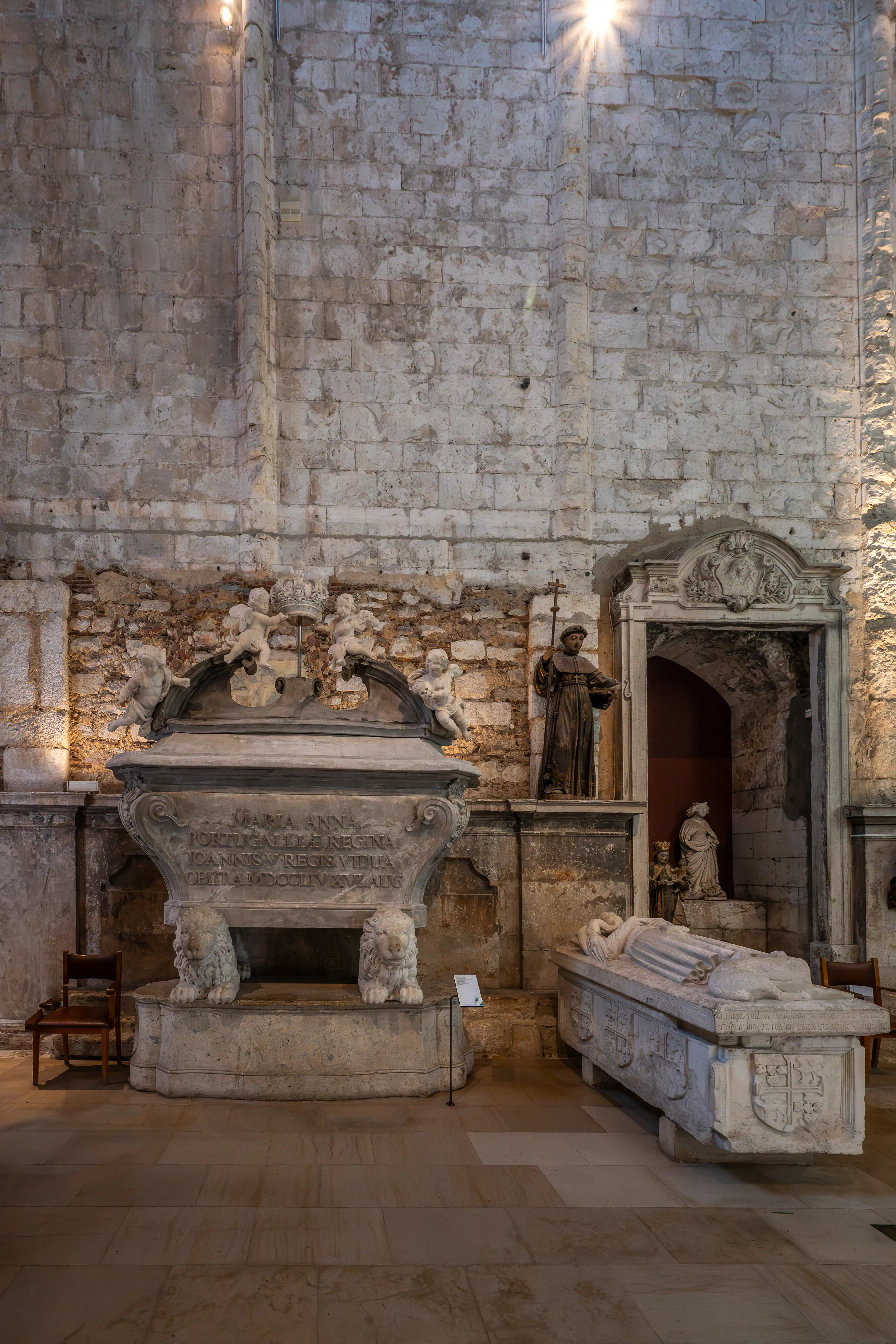 Inside a dimly lit stone church, a large, ornate tomb with cherubs and lions sits in the foreground, while a sarcophagus with a recumbent effigy lies to the right.