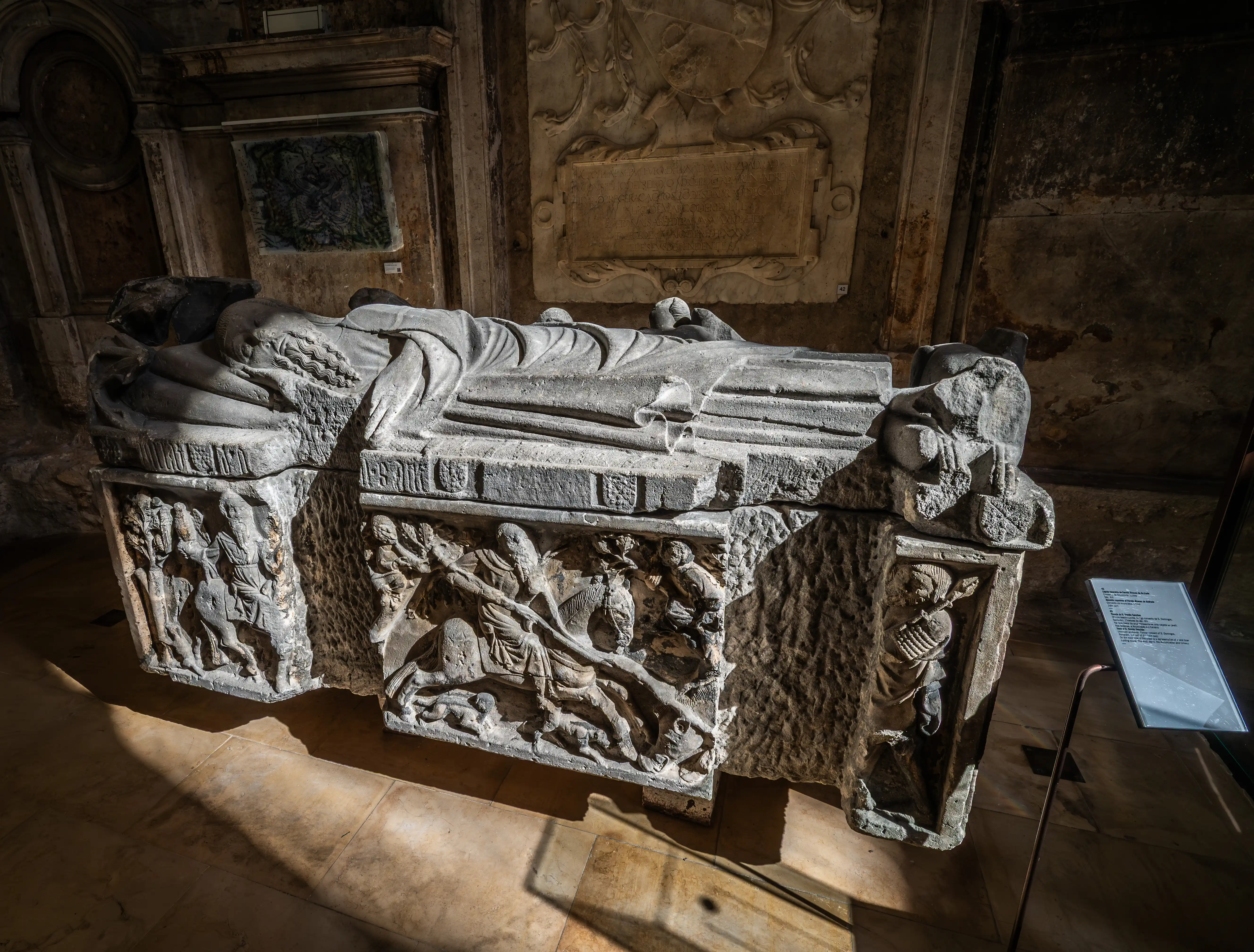  A stone sarcophagus with a sculpted figure lying on top, and carved scenes of riders and warriors on its sides, illuminated by dramatic light and shadow.