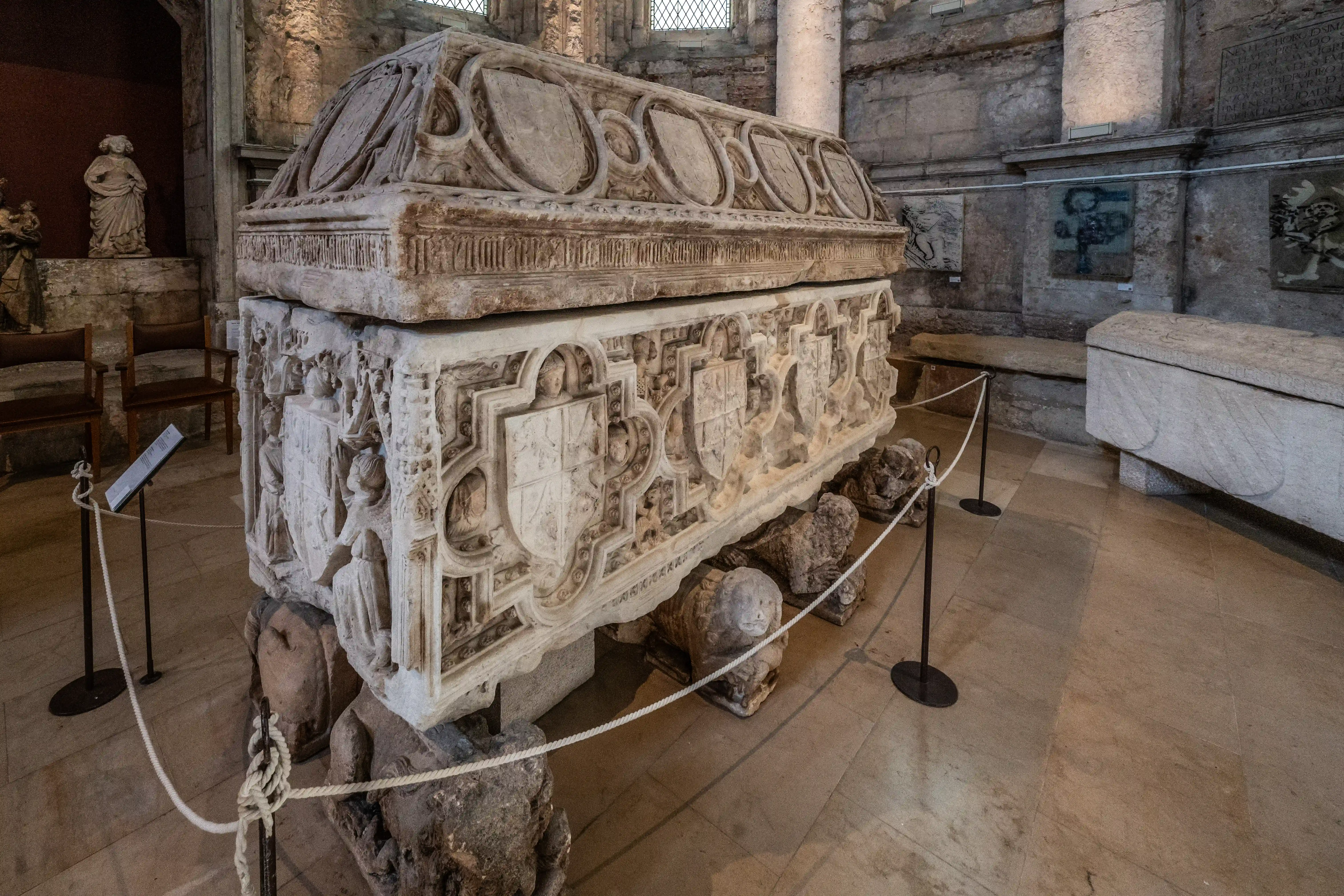A highly detailed, large stone sarcophagus with carved shields and figures, resting on carved lion-like supports, displayed inside a historical stone building.