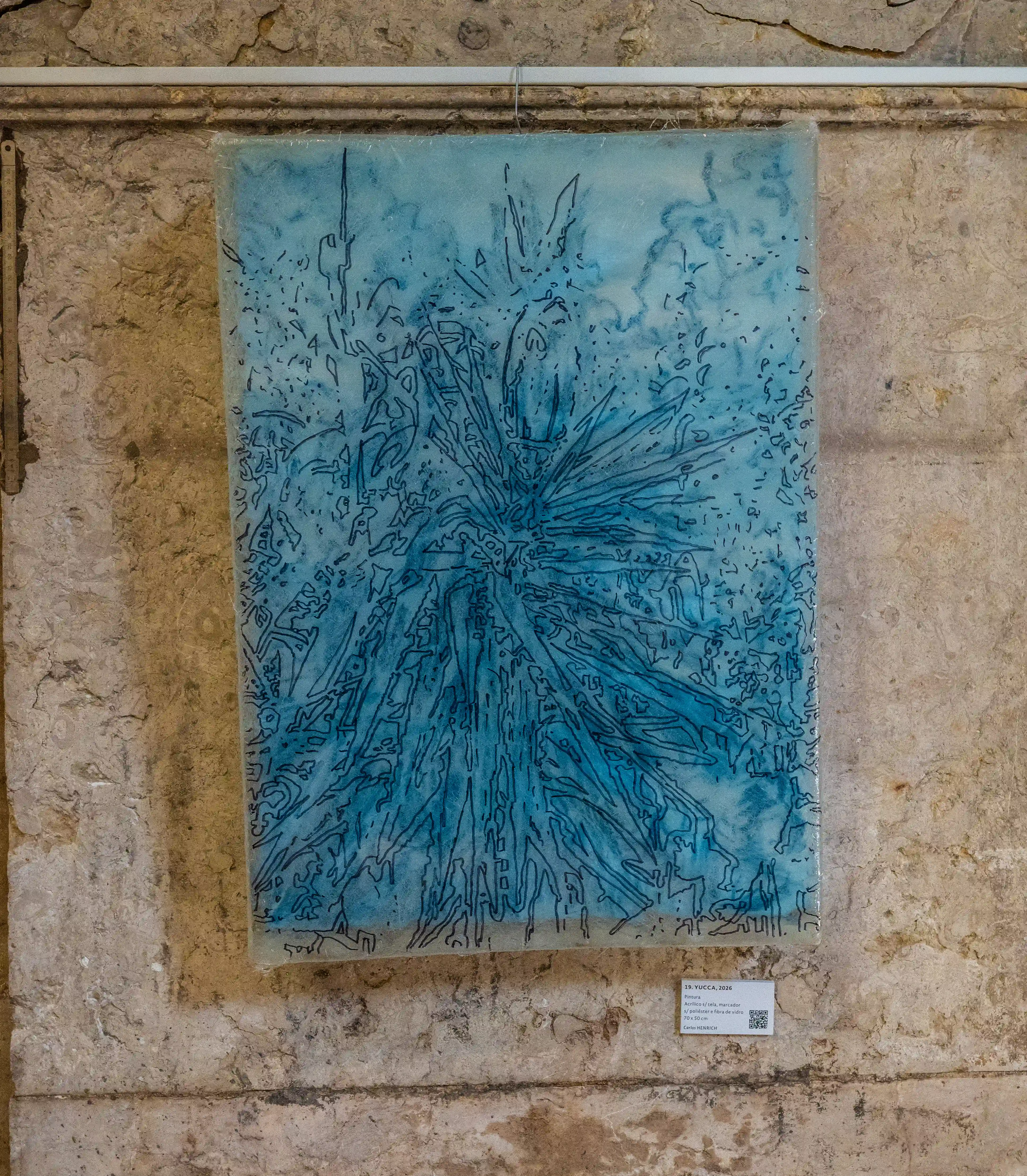 An abstract artwork in shades of blue with dark blue lines, depicting a starburst or plant-like form, is mounted on a rough, textured stone wall. A small label is attached below the artwork.