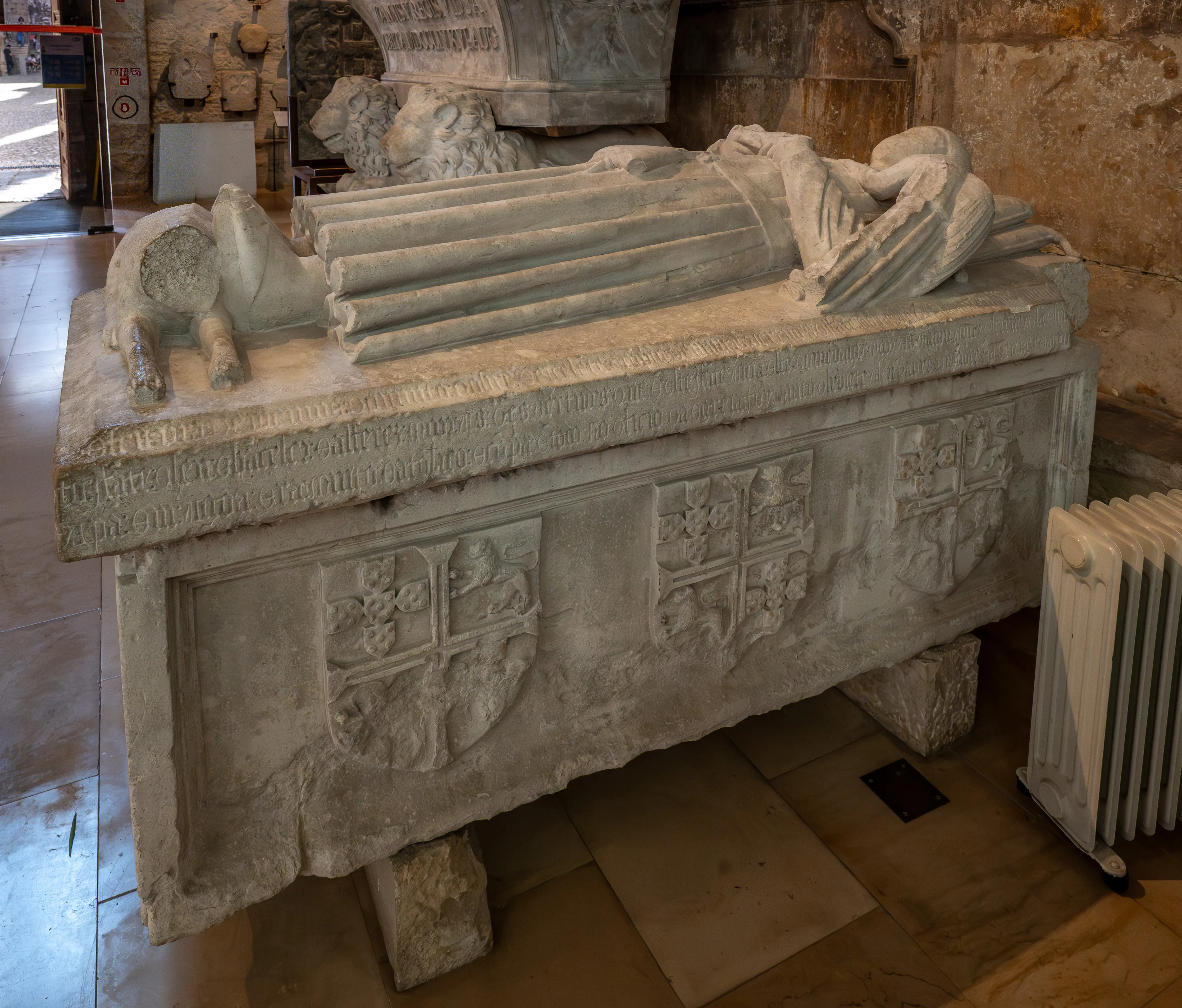 A stone sarcophagus with a carved figure and heraldic shields in a dimly lit historical interior