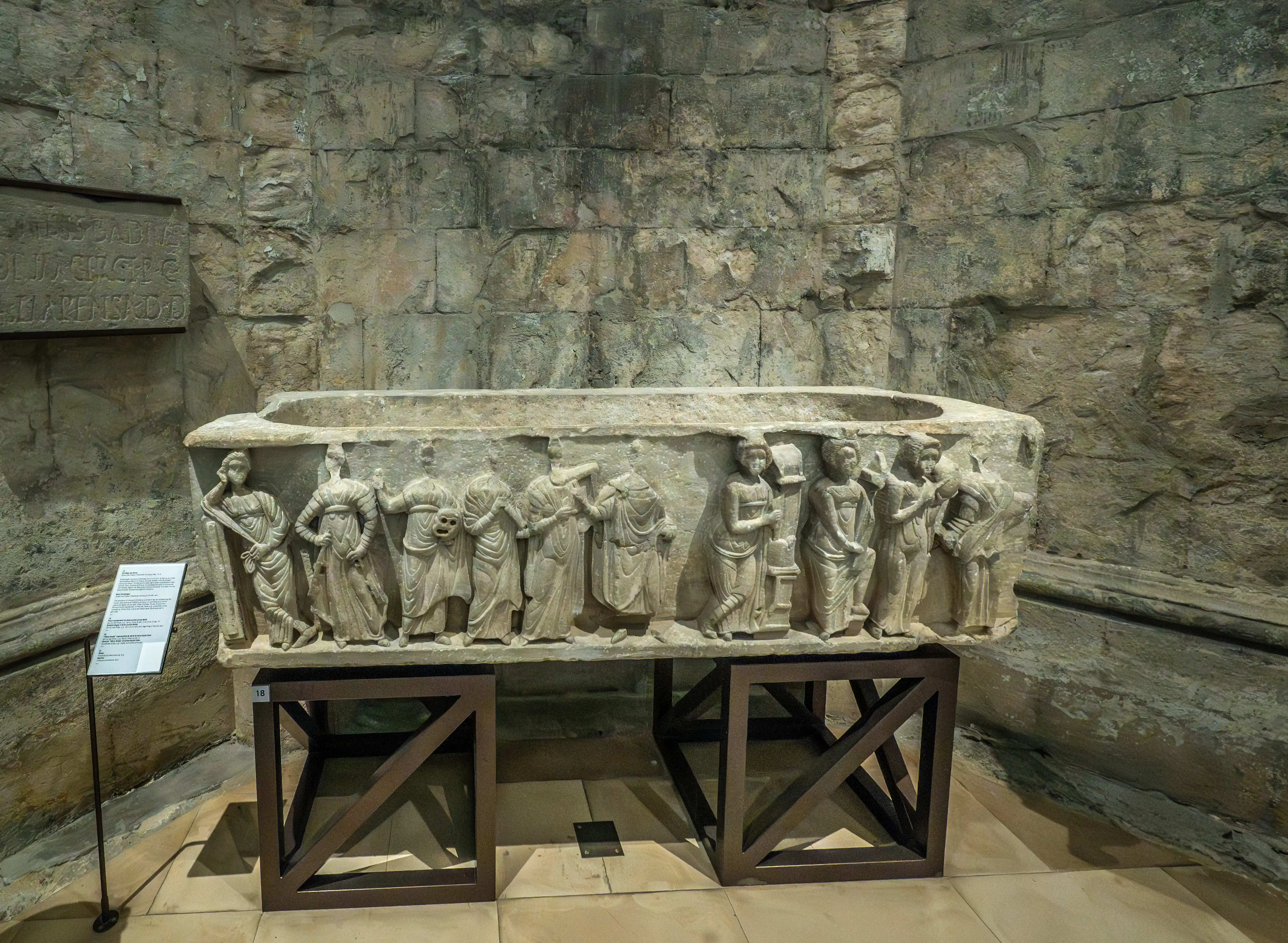 A stone sarcophagus with carved figures and a descriptive plaque, displayed in a stone-walled enclosure.
