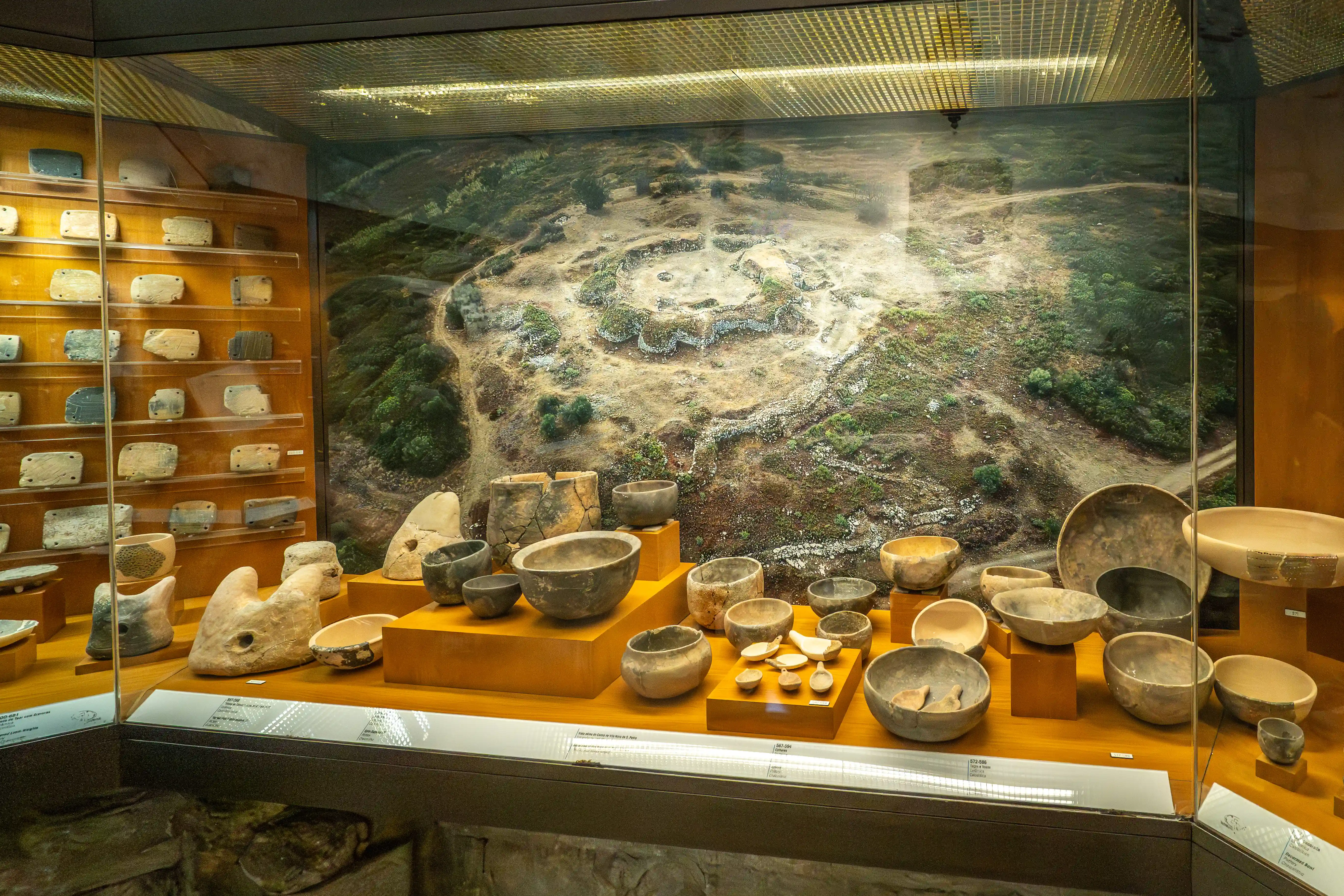 A museum exhibit showcasing numerous ancient ceramic vessels, stone artefacts, and a detailed diorama of an ancient settlement, viewed through a glass display case.