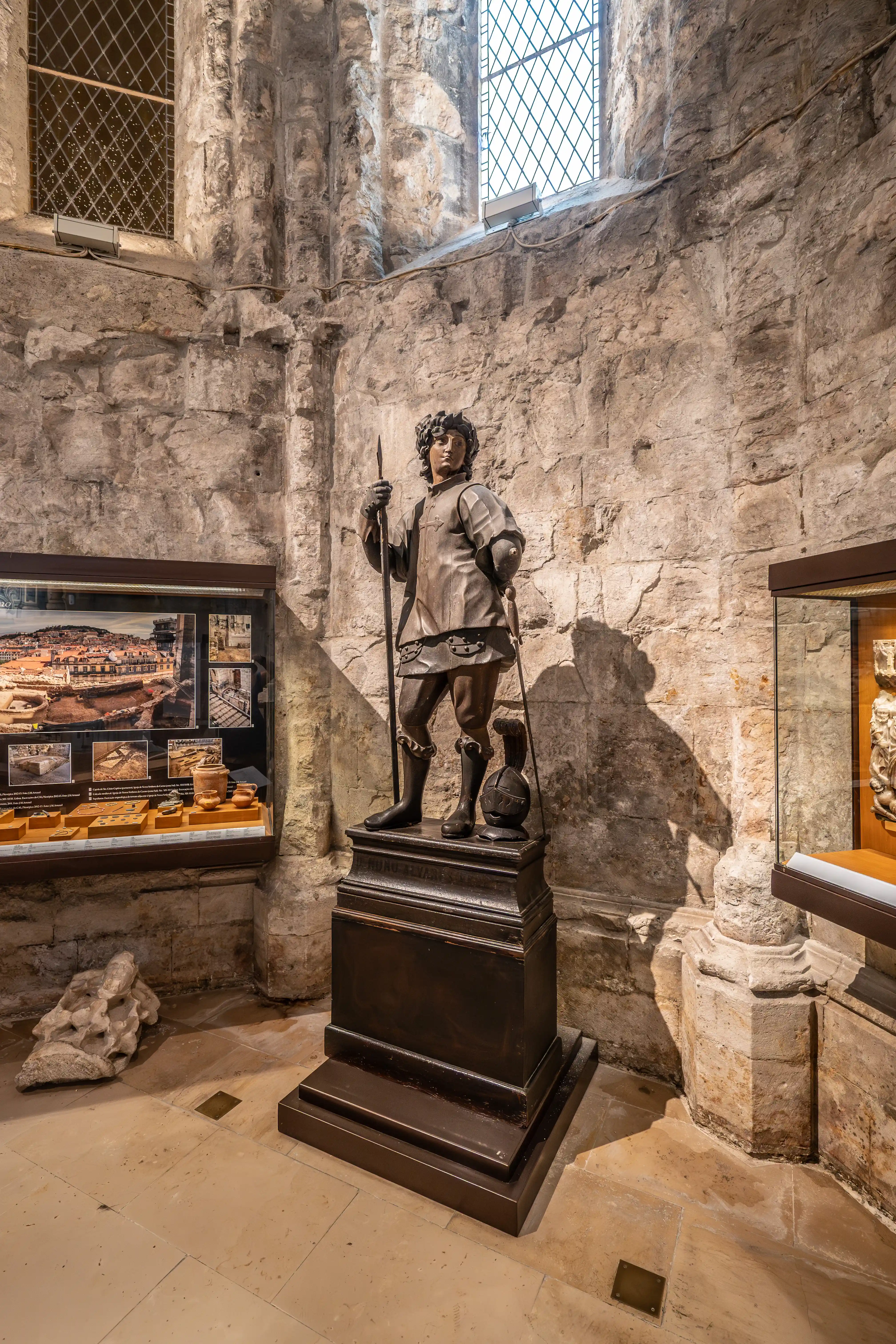 A dark bronze statue of a medieval warrior holding a spear, standing on a dark wooden plinth in a museum. The warrior wears armour and a helmet. Behind the statue is a stone wall with a diamond-paned window. To the left is a display case with artefacts and images, and to the right is another display case with stone carvings.