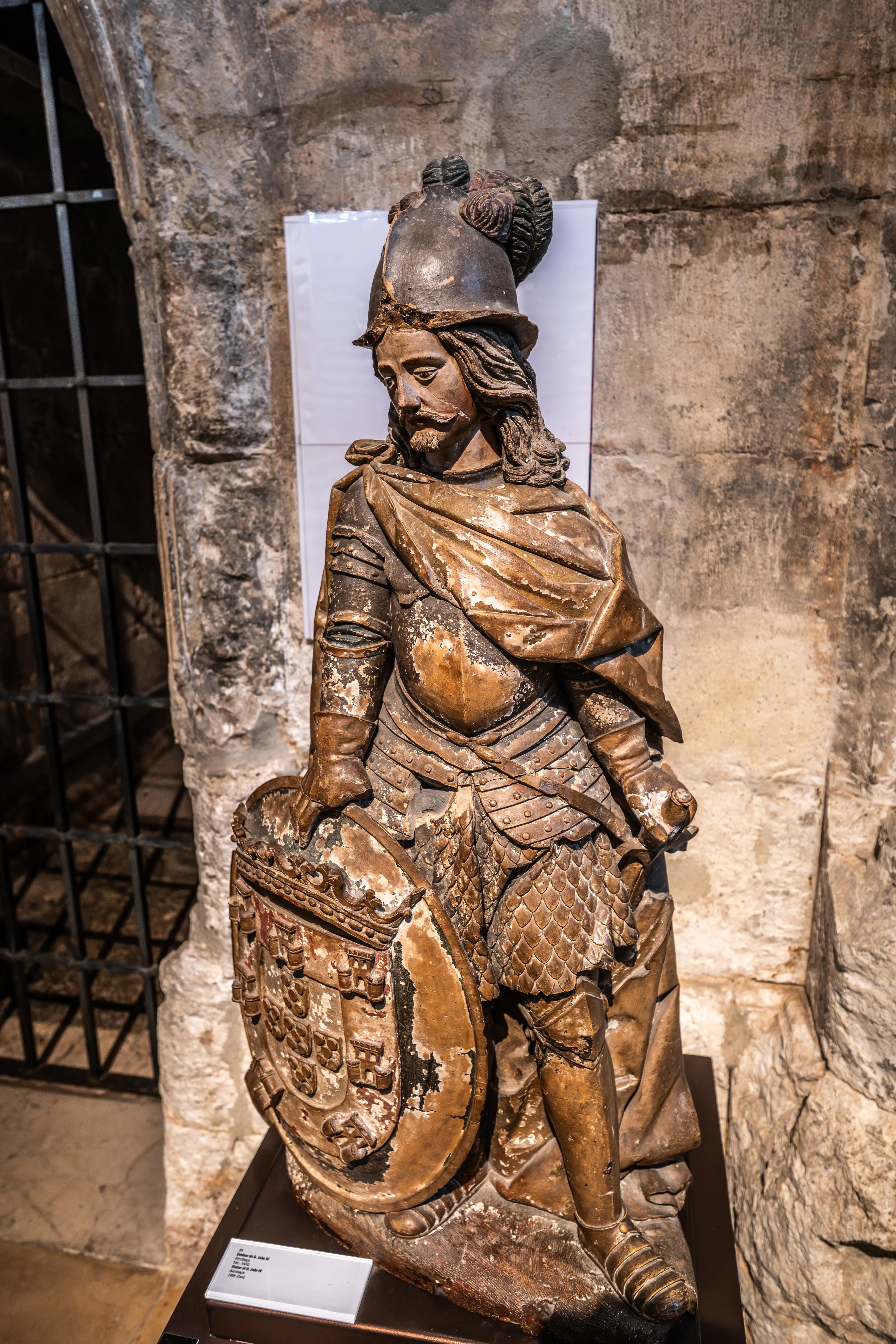 A carved stone statue of a historical figure, likely a nobleman or military leader, standing stoically with a prominent shield in his left hand. He wears a plumed helmet, segmented armour, and a cloak draped over his shoulders. The statue exhibits signs of age and wear