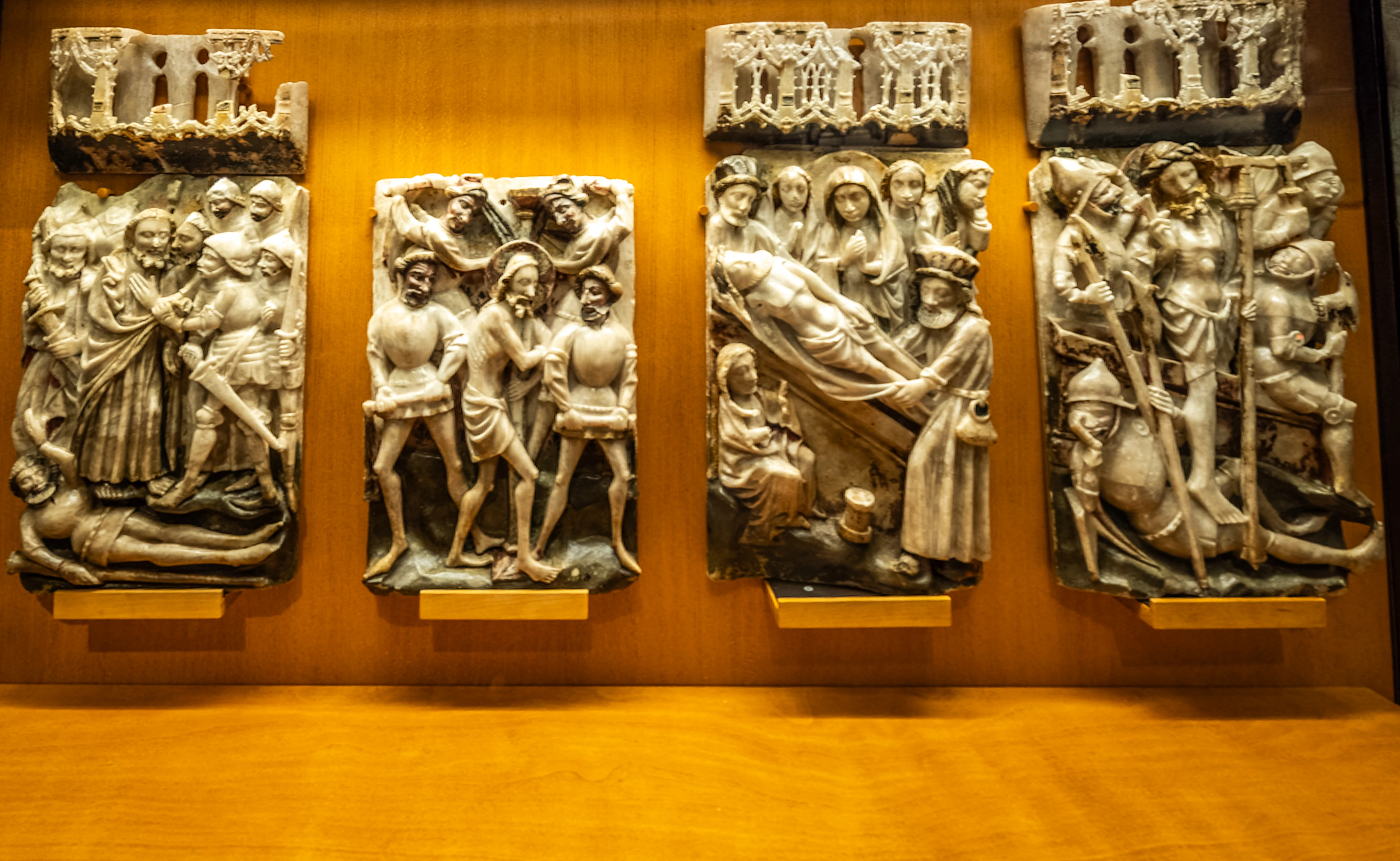 Four framed relief carvings, carved from light-coloured material, are displayed on a wooden shelf in a museum. Each relief depicts a religious scene with multiple figures in a historical style. The scenes appear to represent moments from the Passion and Resurrection of Christ, including an arrest, a scourging, and the entombment and resurrection.