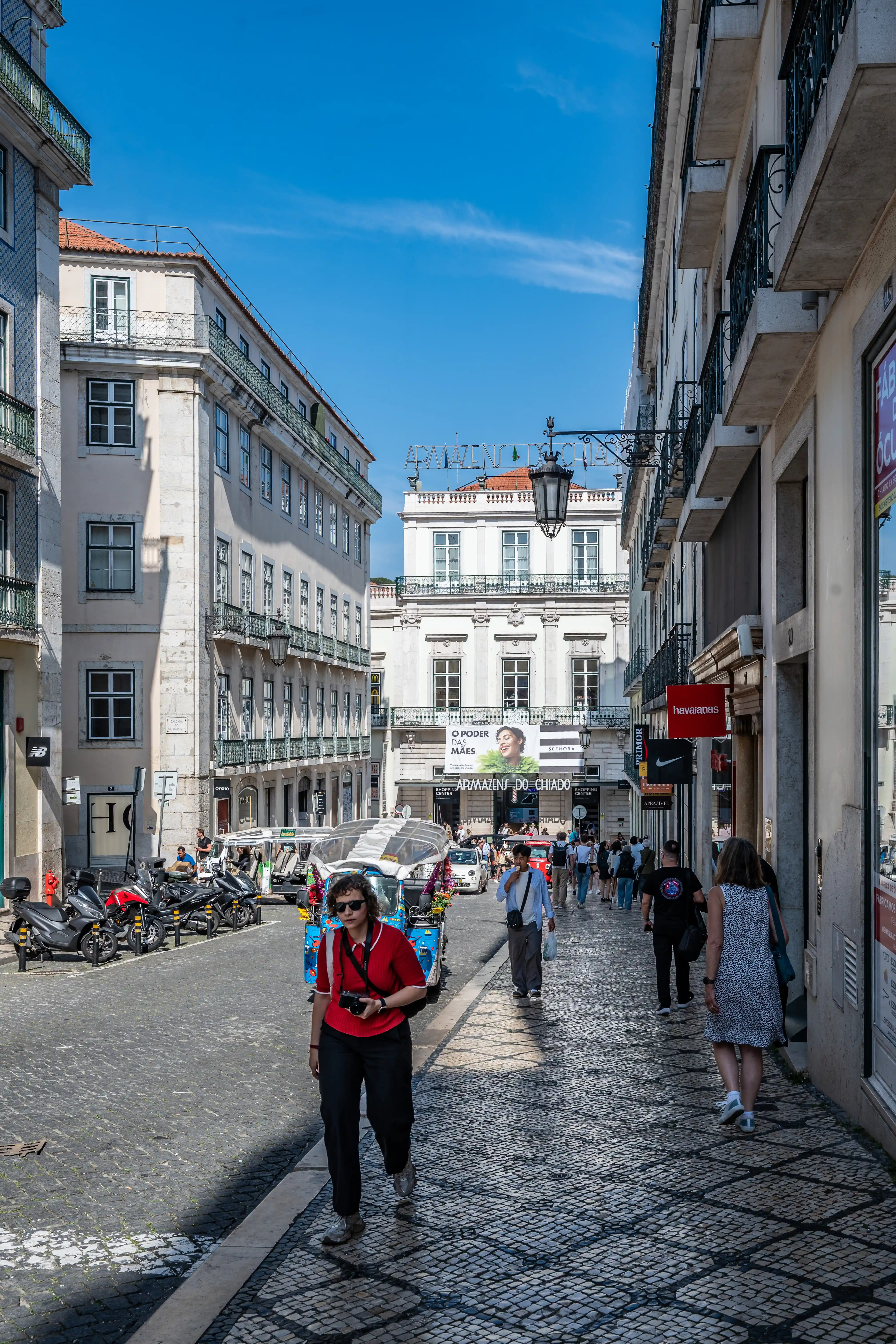 A woman in a red top and black trousers walks down a cobblestone street in Lisbon, holding a camera. Buildings with balconies line the street, and other pedestrians are visible. A blue tuk-tuk is parked on the left.