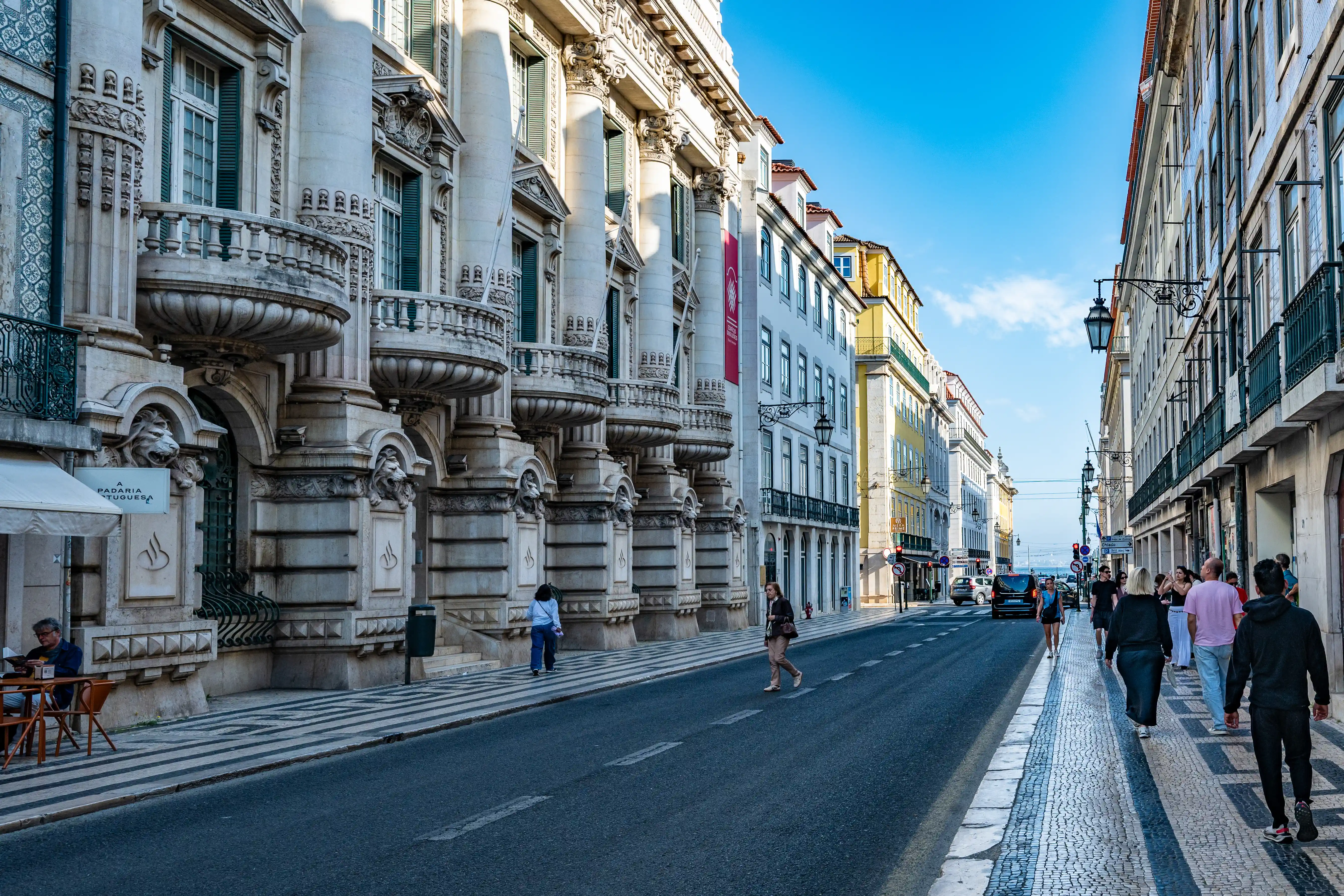 A wide street in Lisbon with ornate classical buildings on the left and more modern structures on the right. People are walking on the patterned sidewalk on the right side of the street. Cars are visible on the road.