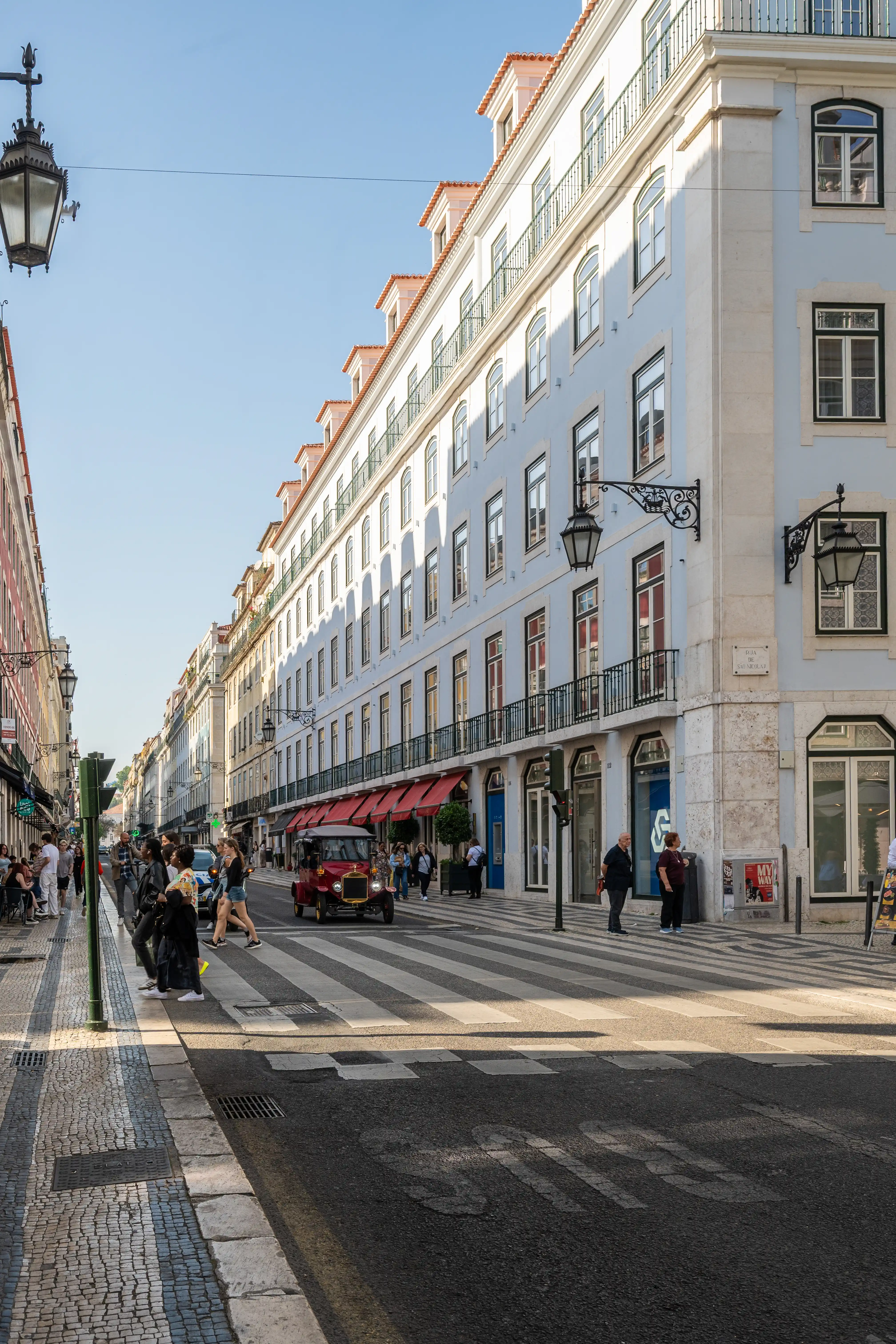 A wide street in Lisbon with pale blue buildings, wrought-iron balconies, and ornate street lamps. A vintage red car is crossing a zebra crossing on a sunny day, with several pedestrians on the pavemen