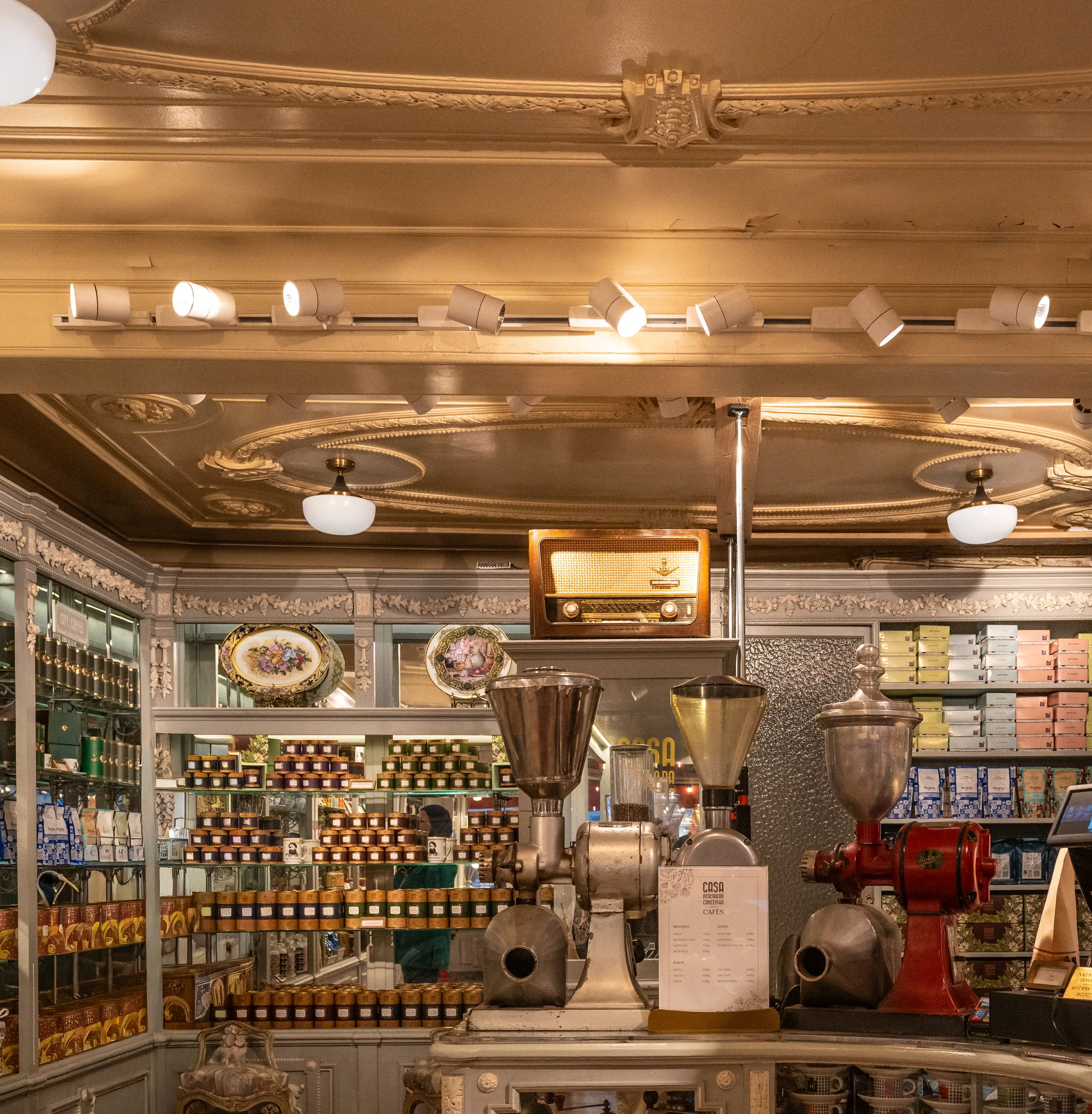 Interior of a vintage tea and coffee shop with shelves stocked with colourful tins. A woman is seen from behind, looking at the products. Vintage coffee grinders and a retro radio are displayed on the counter.