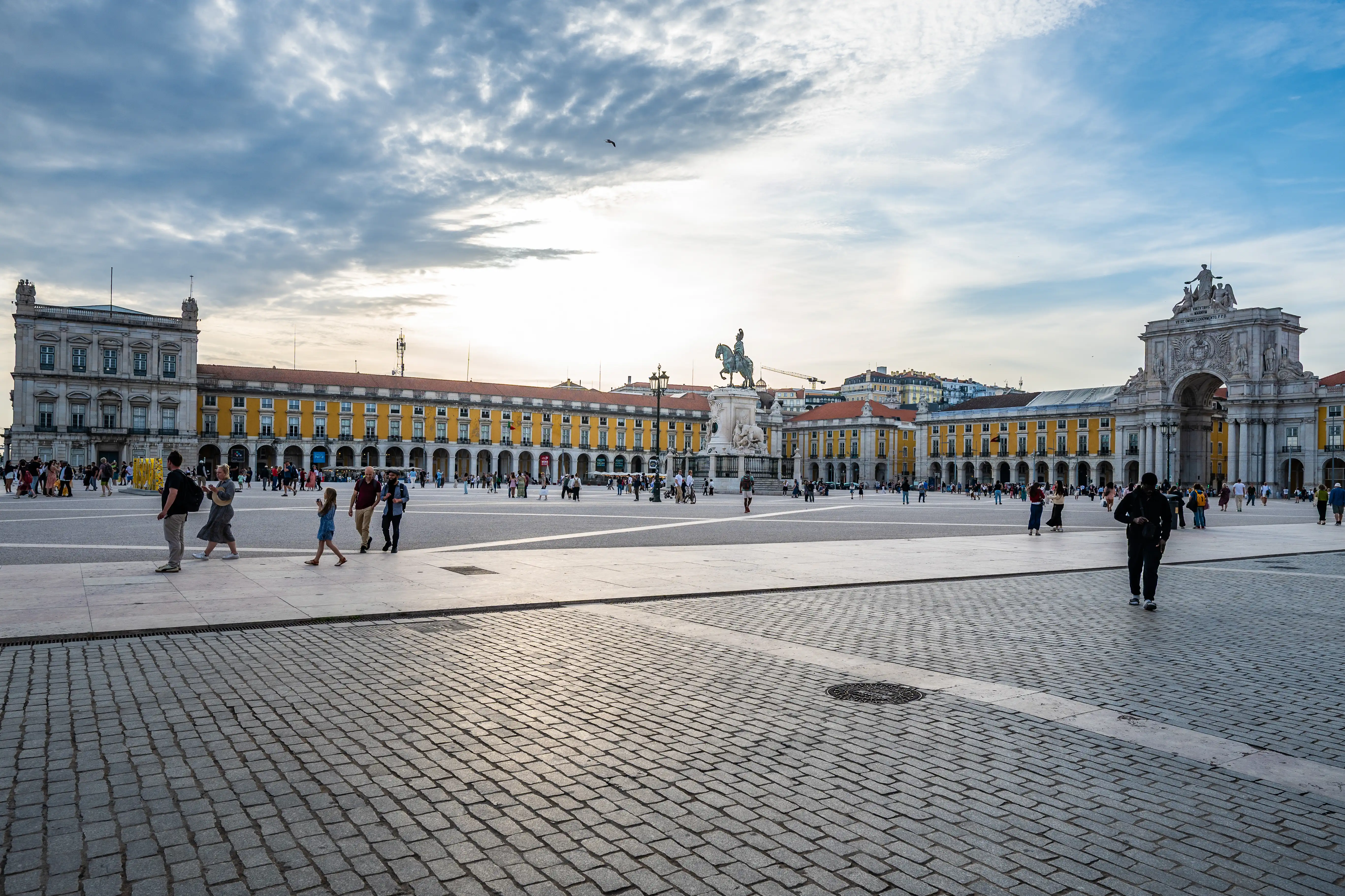 A spacious, historic European square paved with cobblestones, featuring large, ornate buildings with yellow facades and arcades, a statue of a man on horseback, and a triumphal arch. Several people are walking in the open space.