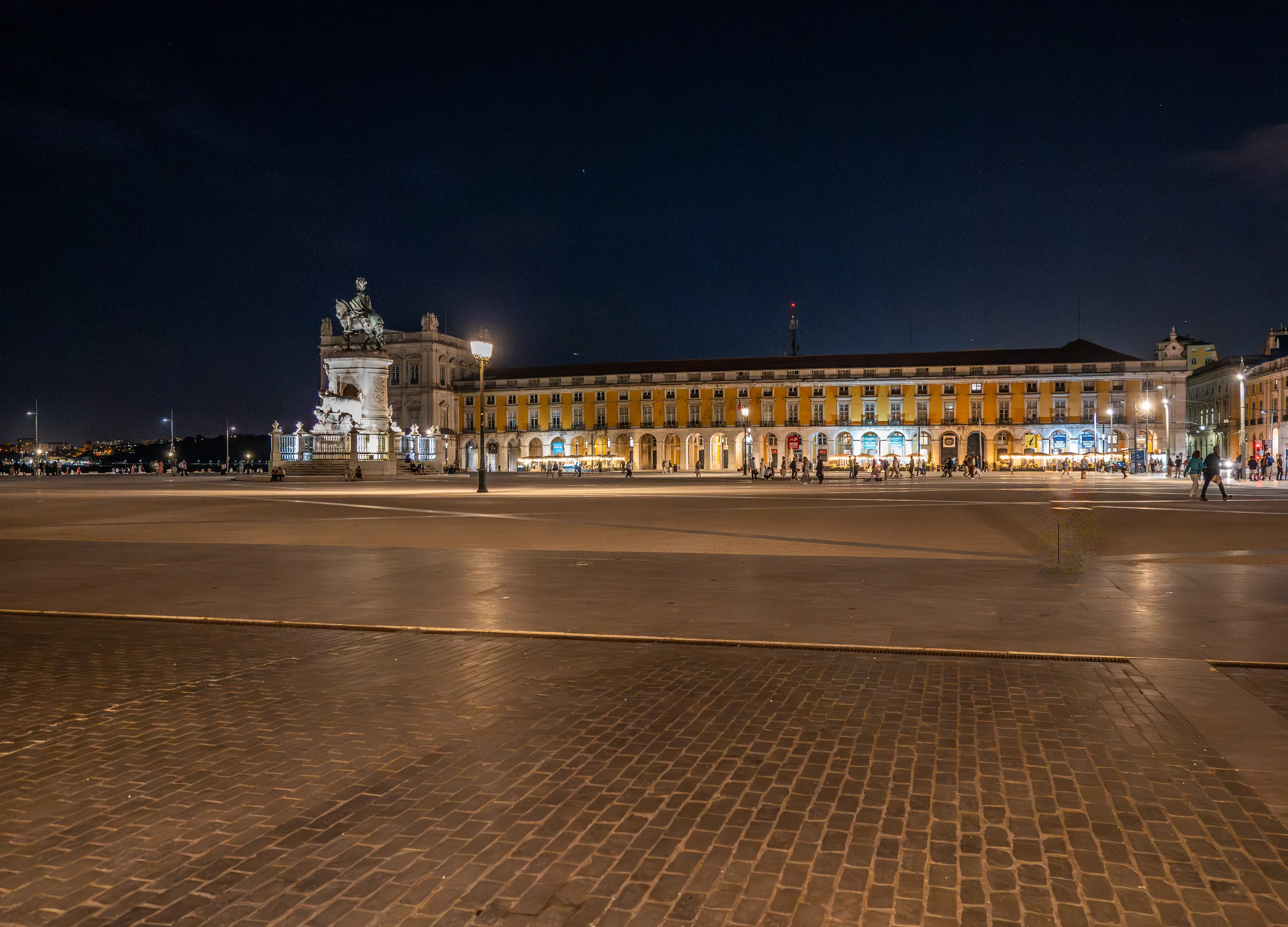 Praça do Comércio in Lisbon at night, showing illuminated buildings, an equestrian statue, and people walking on the square.