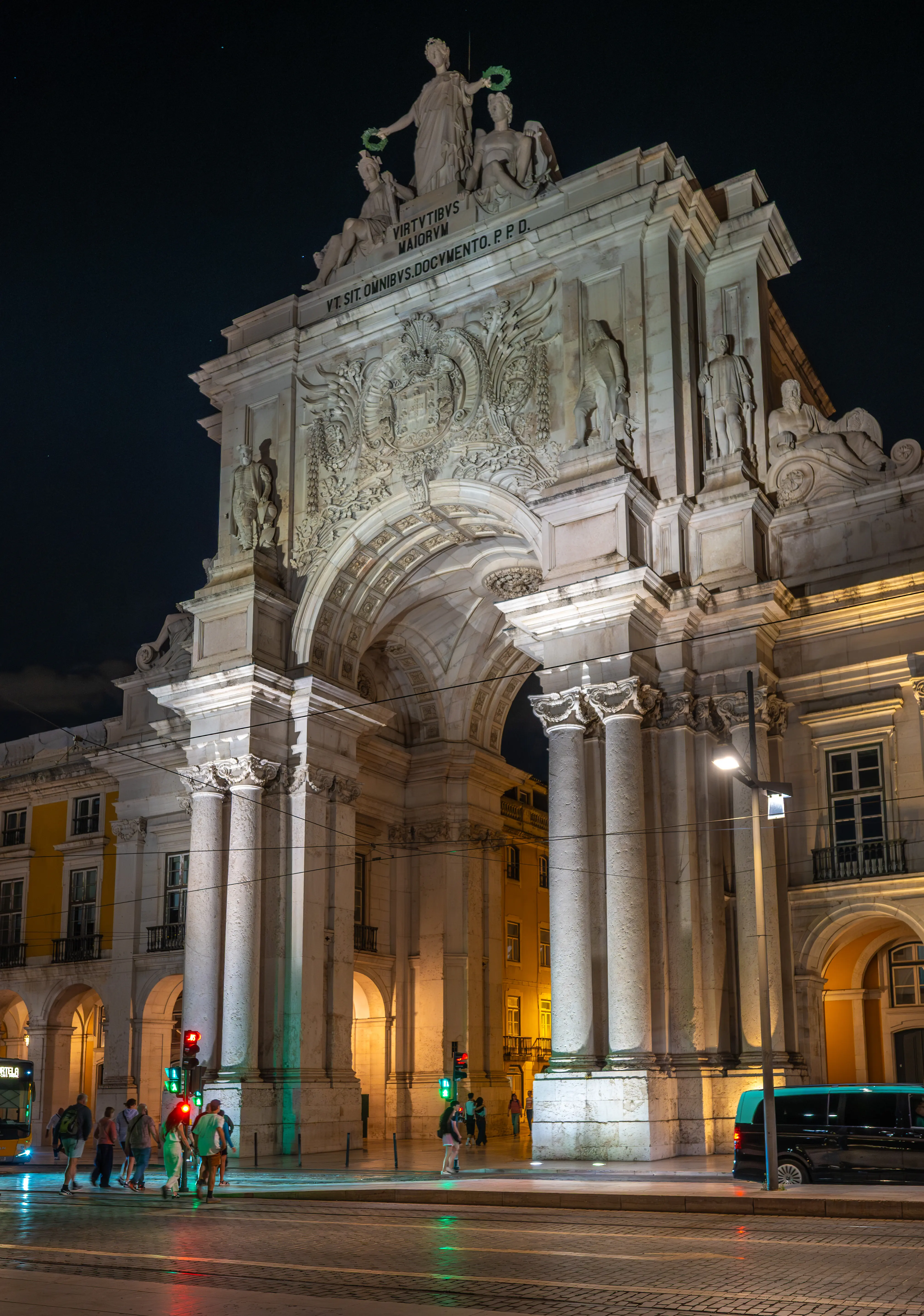 Night view of Praça do Comércio in Lisbon, Portugal, featuring the illuminated Arco da Rua Augusta and yellow buildings with arcades, a bus, and pedestrians.