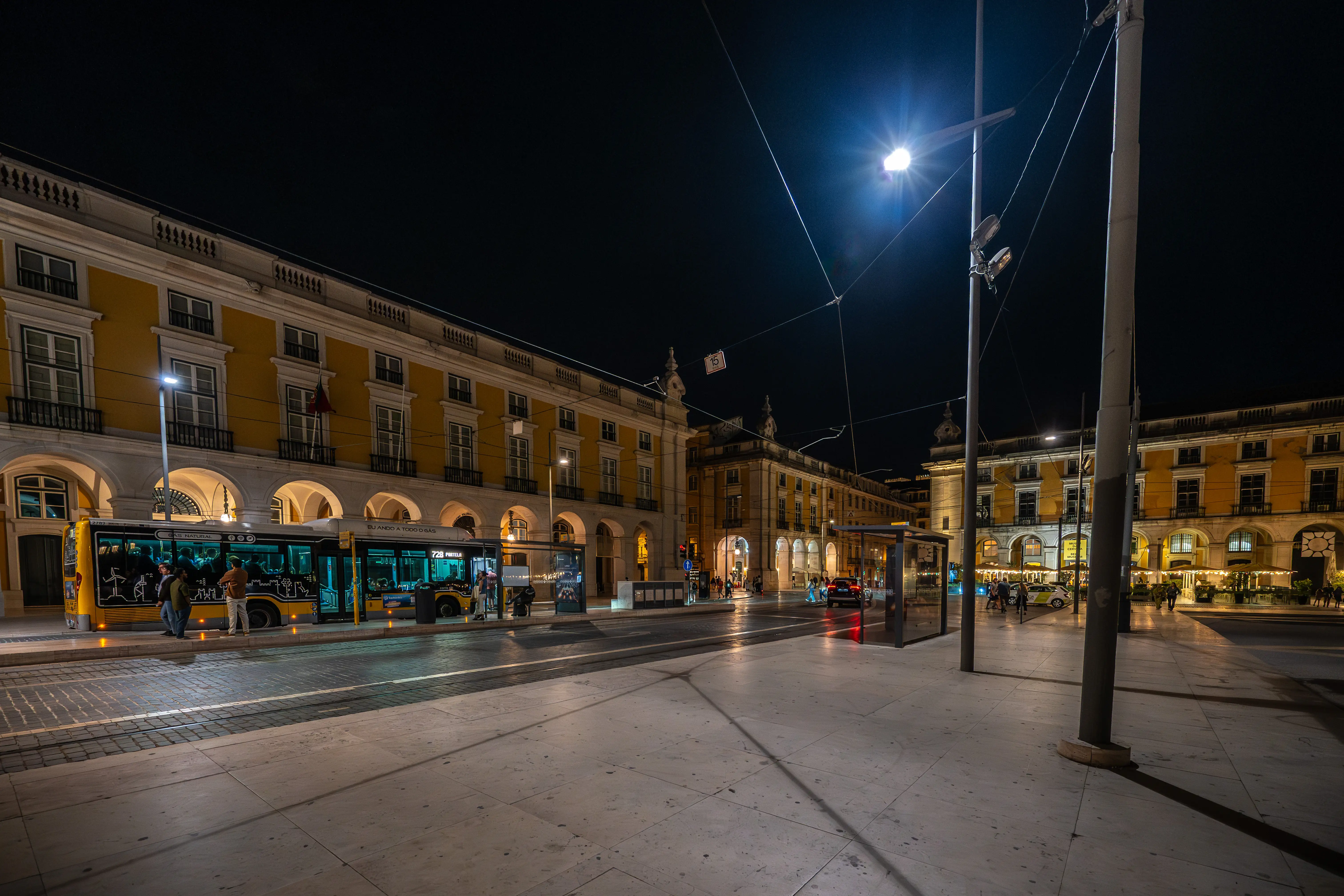 Praça do Comércio, Lisbon, at night, showing yellow buildings with arcades, a tram, streetlights, and paved ground.
