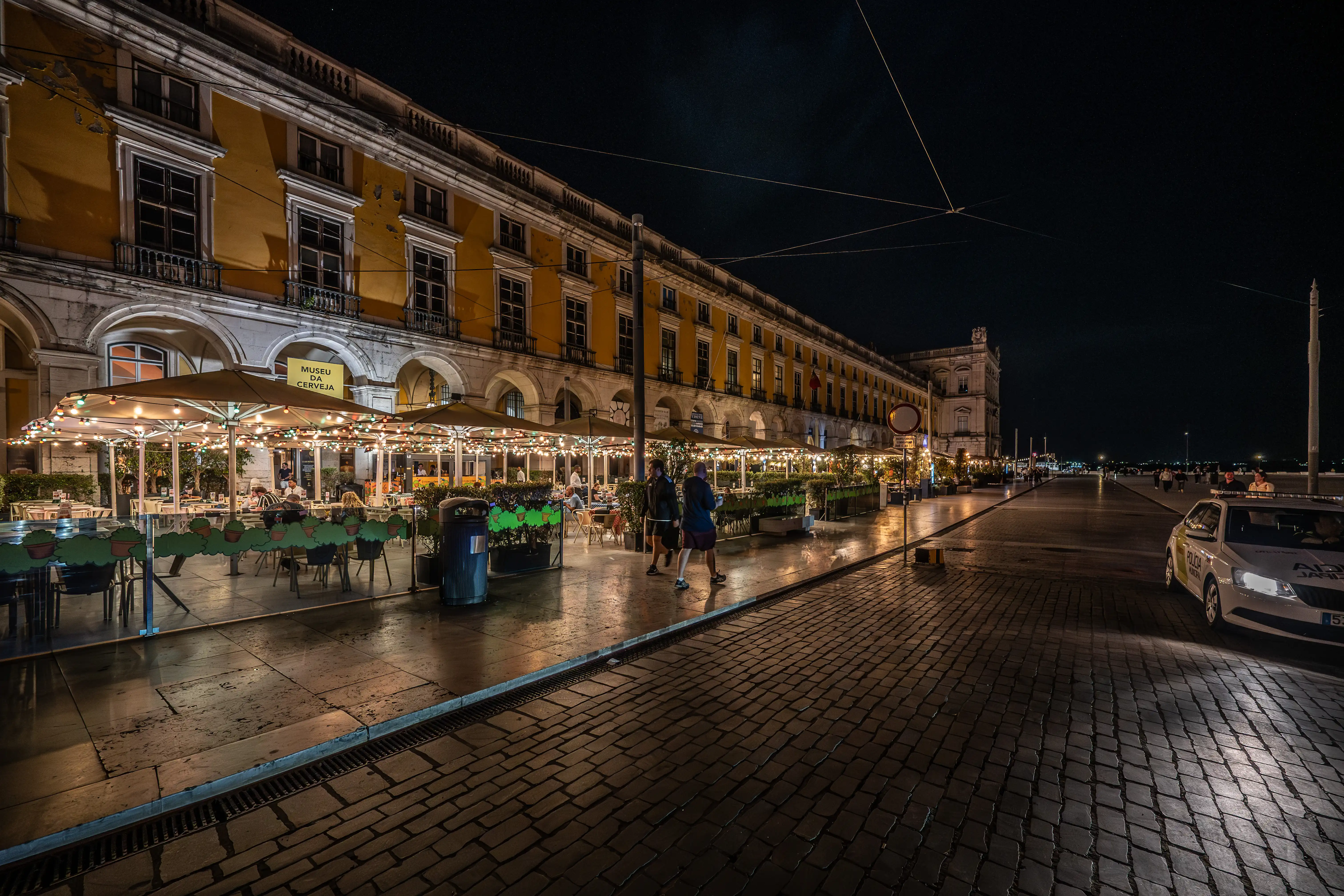 A Lisbon square at night, showing a long yellow building with arched walkways housing an outdoor cafe, with cobblestones and a police car in the foreground.