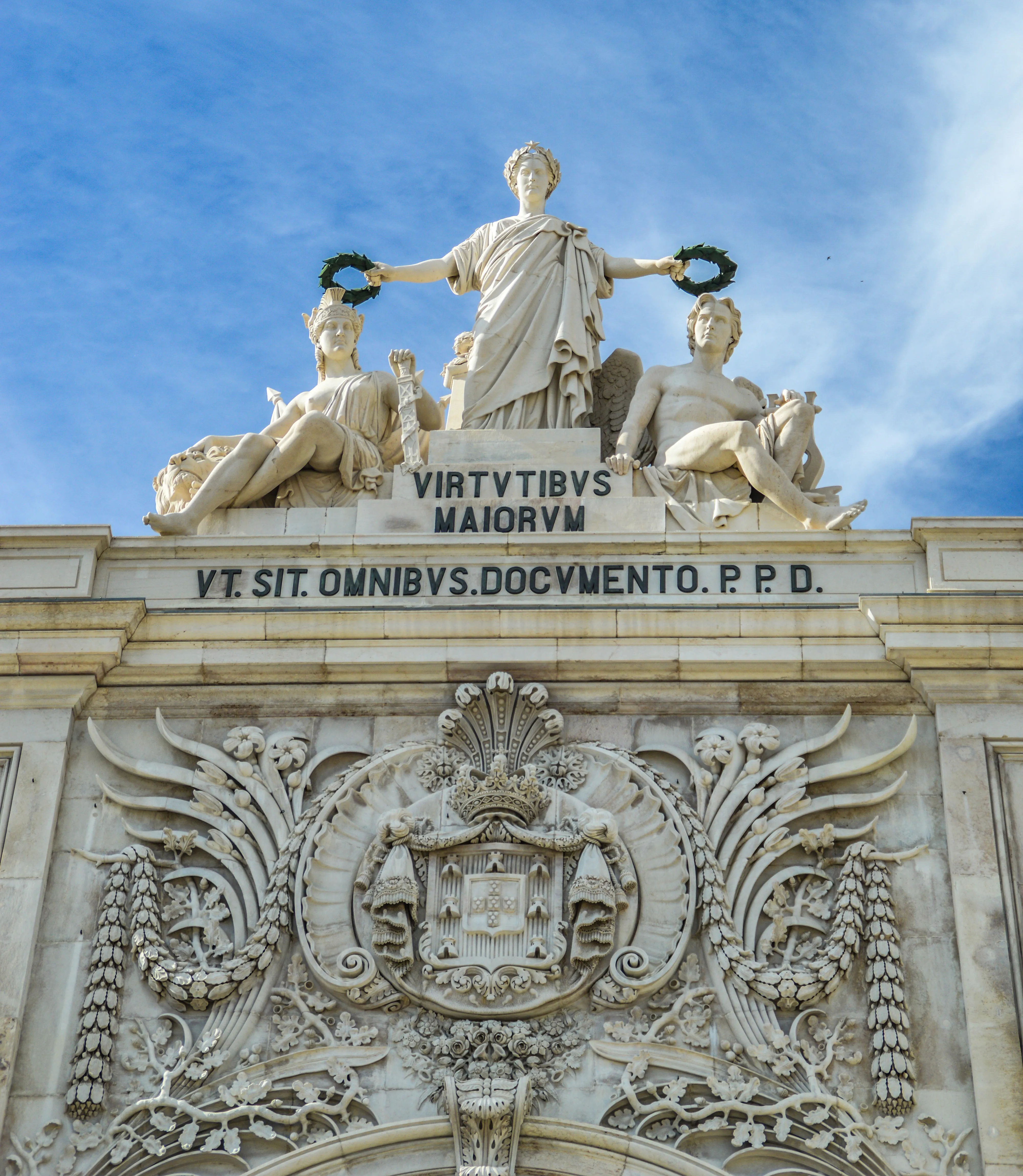 An ornate white marble sculpture of three allegorical figures crowning the top of an archway. A central female figure holds wreaths in each hand, while a seated female figure to her left holds a sword and a seated male figure to her right has an angelic wing. Below the sculptures are two Latin inscriptions and elaborate floral and crest carvings.