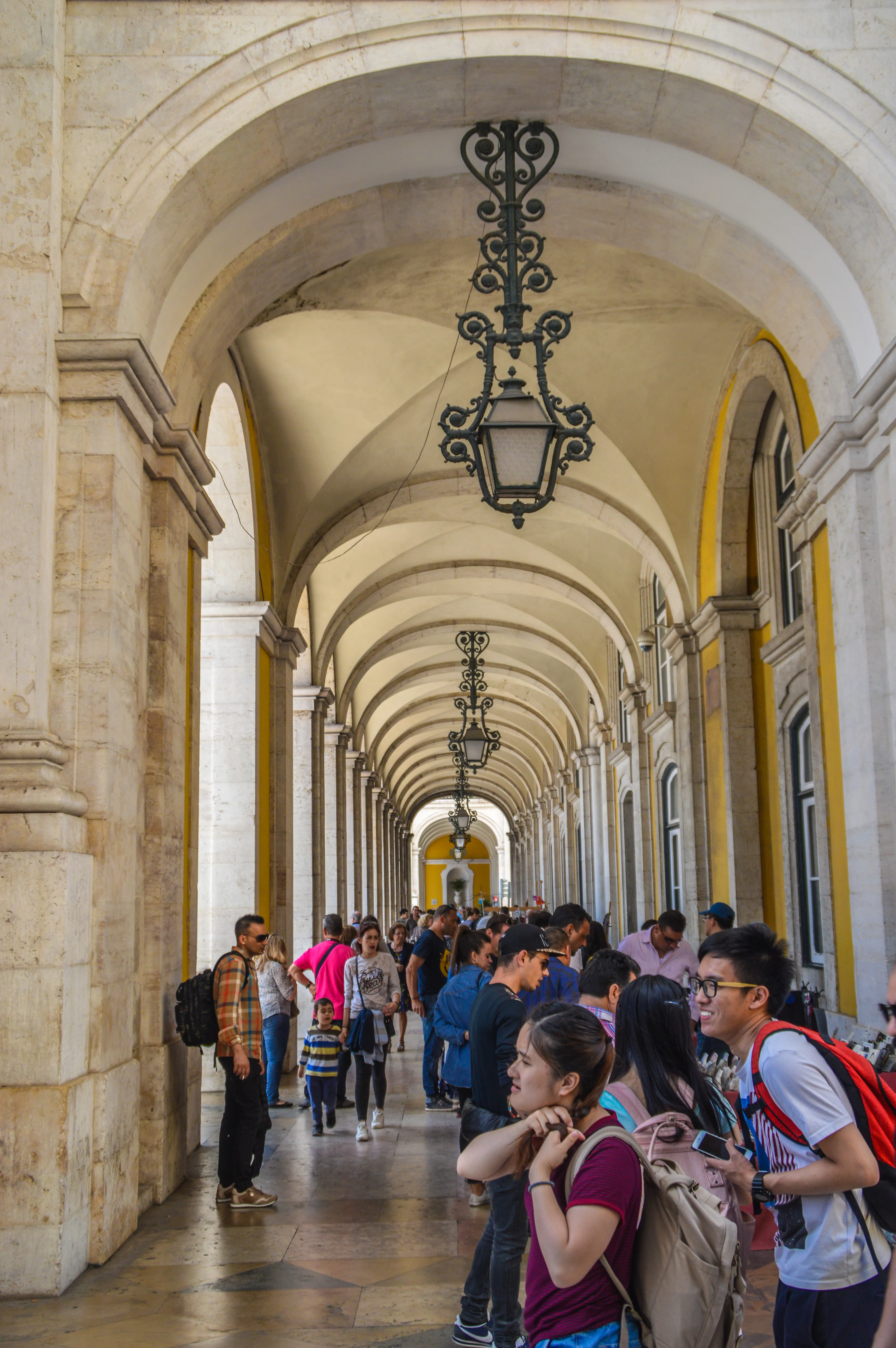 People walking through a long, covered arcade with arched stone ceilings and ornate hanging lamps.