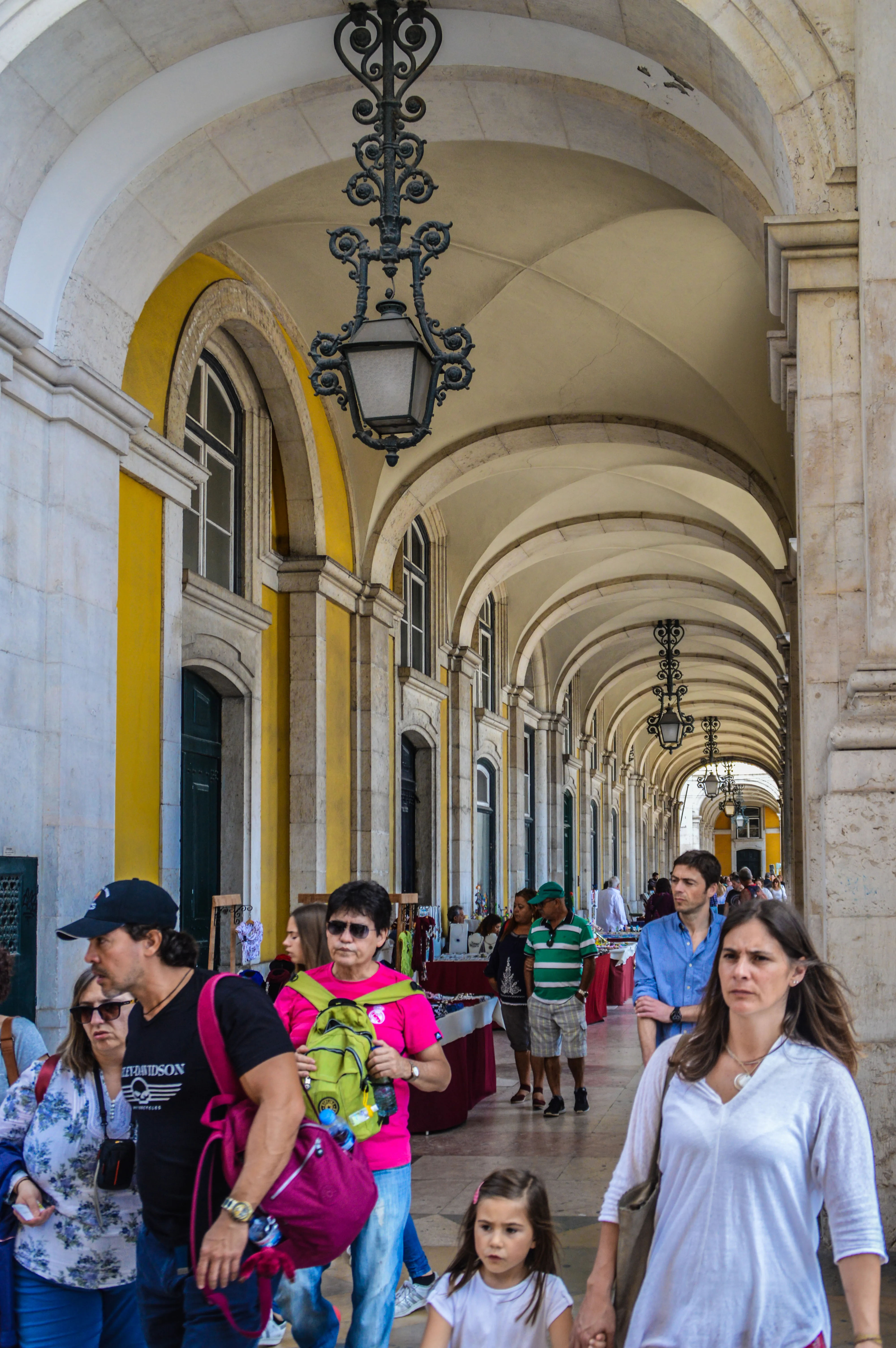 A diverse group of people walk through a sunlit, yellow-walled arcade lined with arched openings and decorative hanging lanterns. Some individuals are looking at market stalls set up along the walkway.