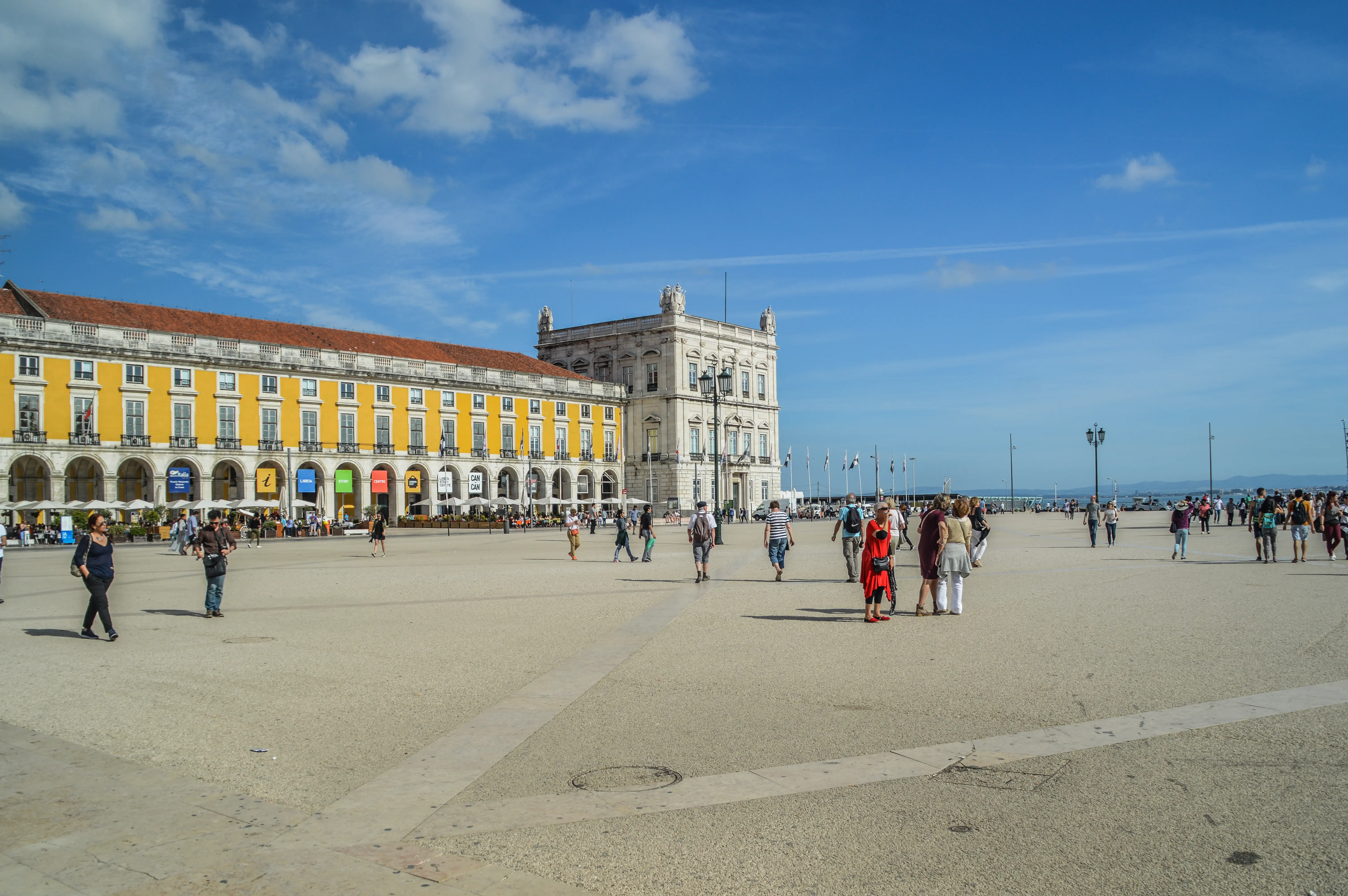 People walking in a large, open square in front of a yellow building with arched walkways under a blue sky.