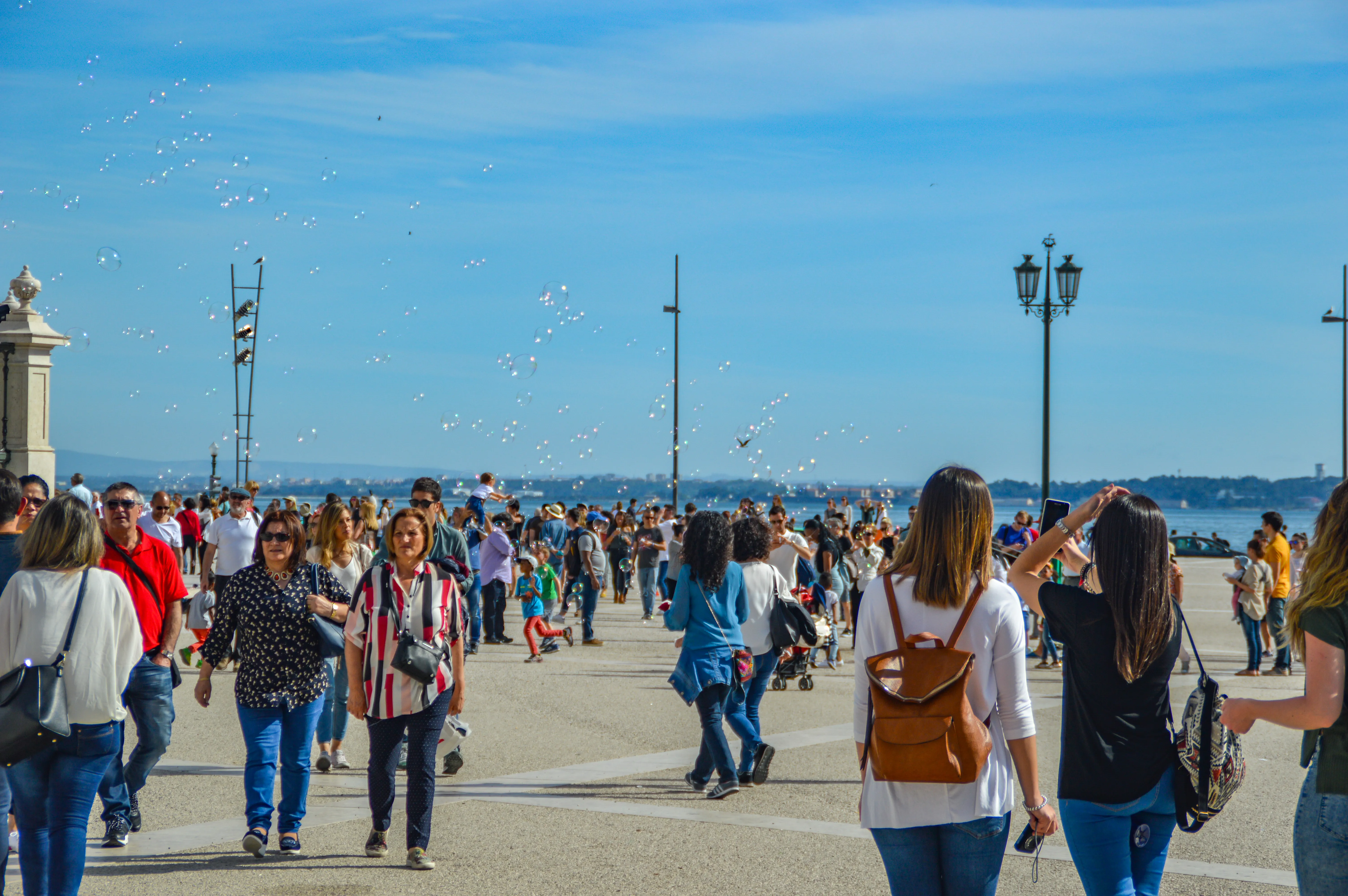 A wide shot of a sunny promenade filled with people walking and small iridescent bubbles floating in the blue sky.