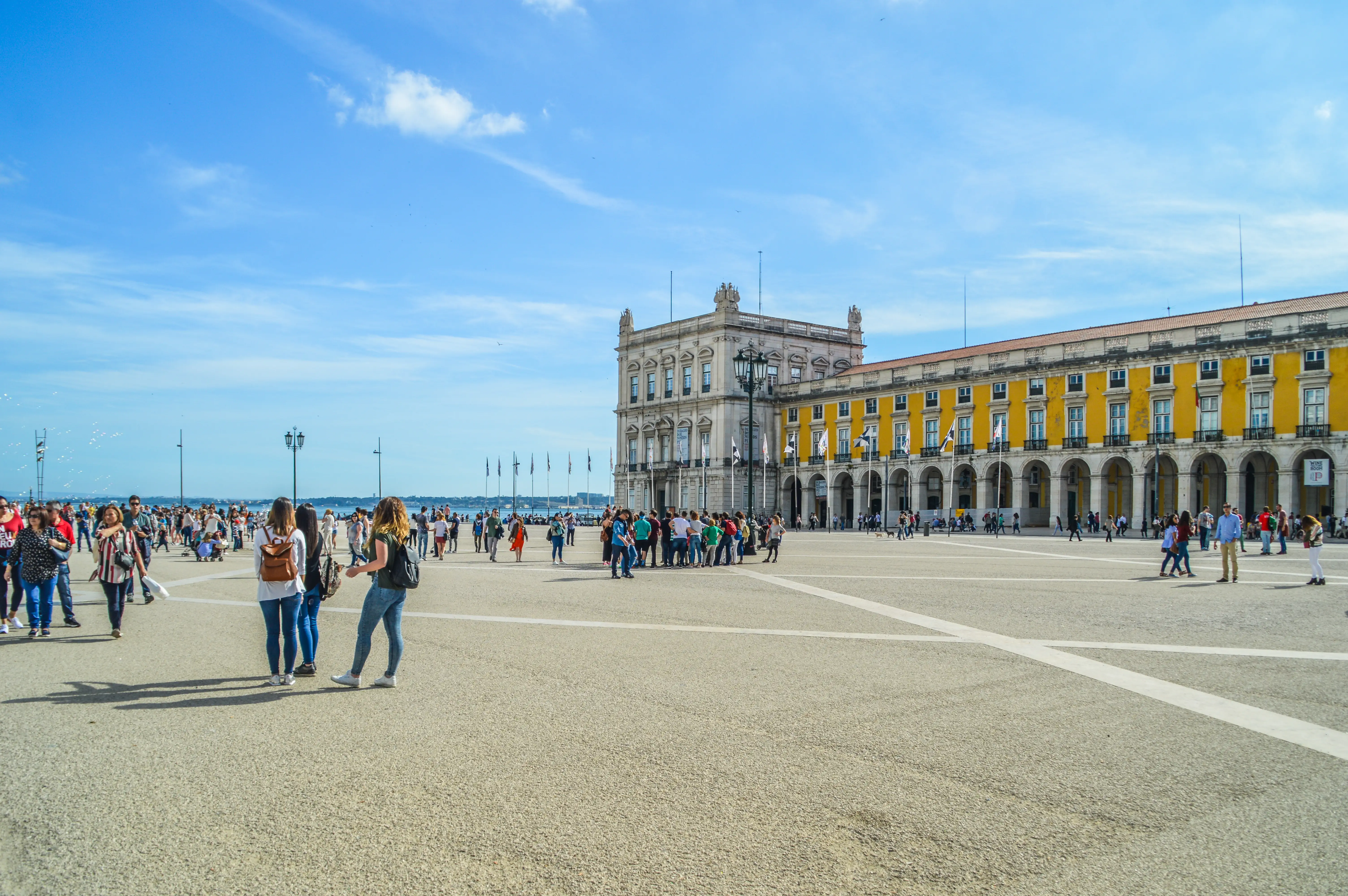 People stroll across the expansive, sunlit Praça do Comércio in Lisbon, with the grand yellow and white historic buildings forming a backdrop under a bright blue sky.