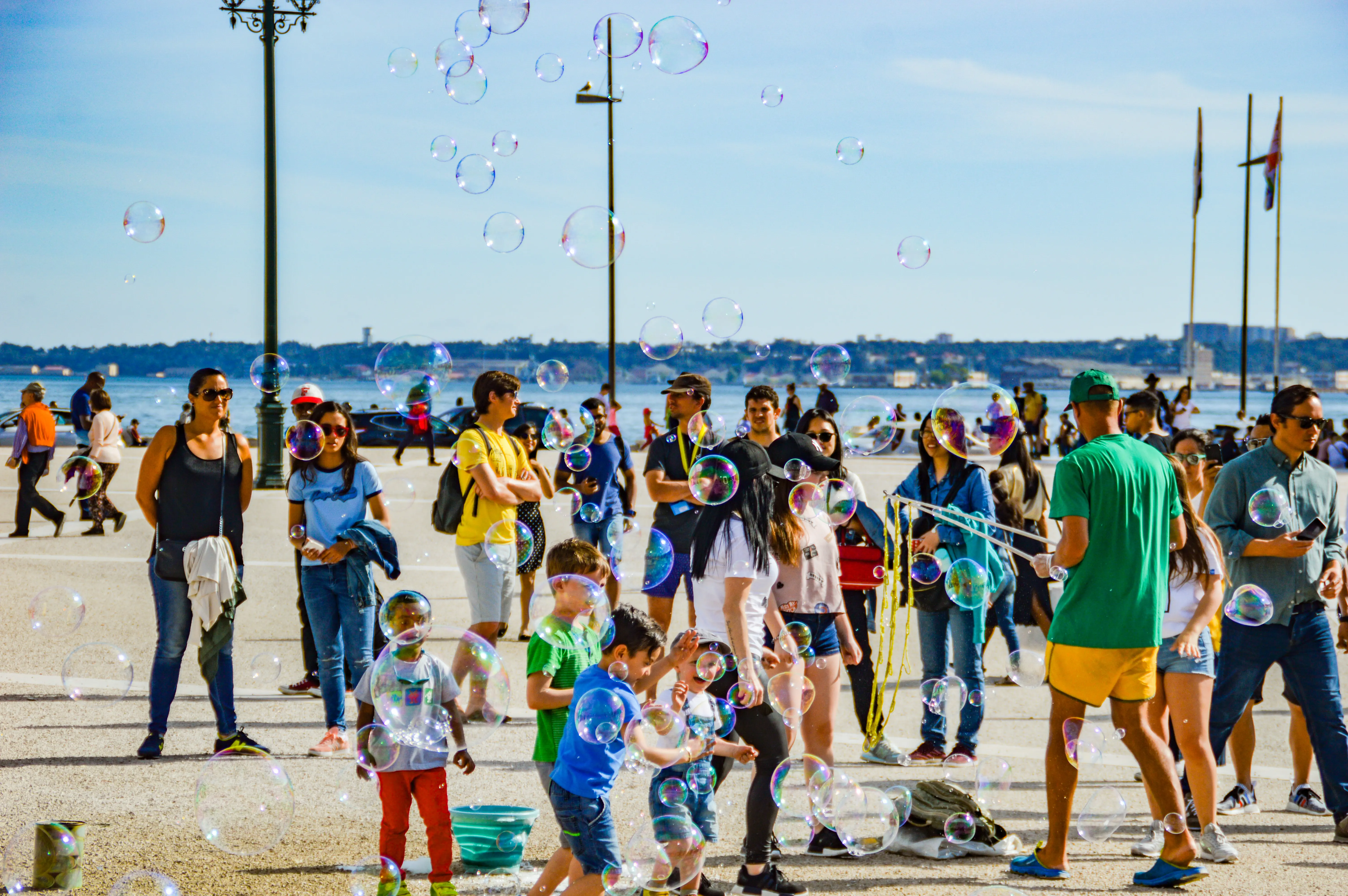 A group of people, including many children, are playing with numerous large, iridescent bubbles floating in the air on a sunny day near a waterfront. A man is blowing bubbles in the centre of the image.